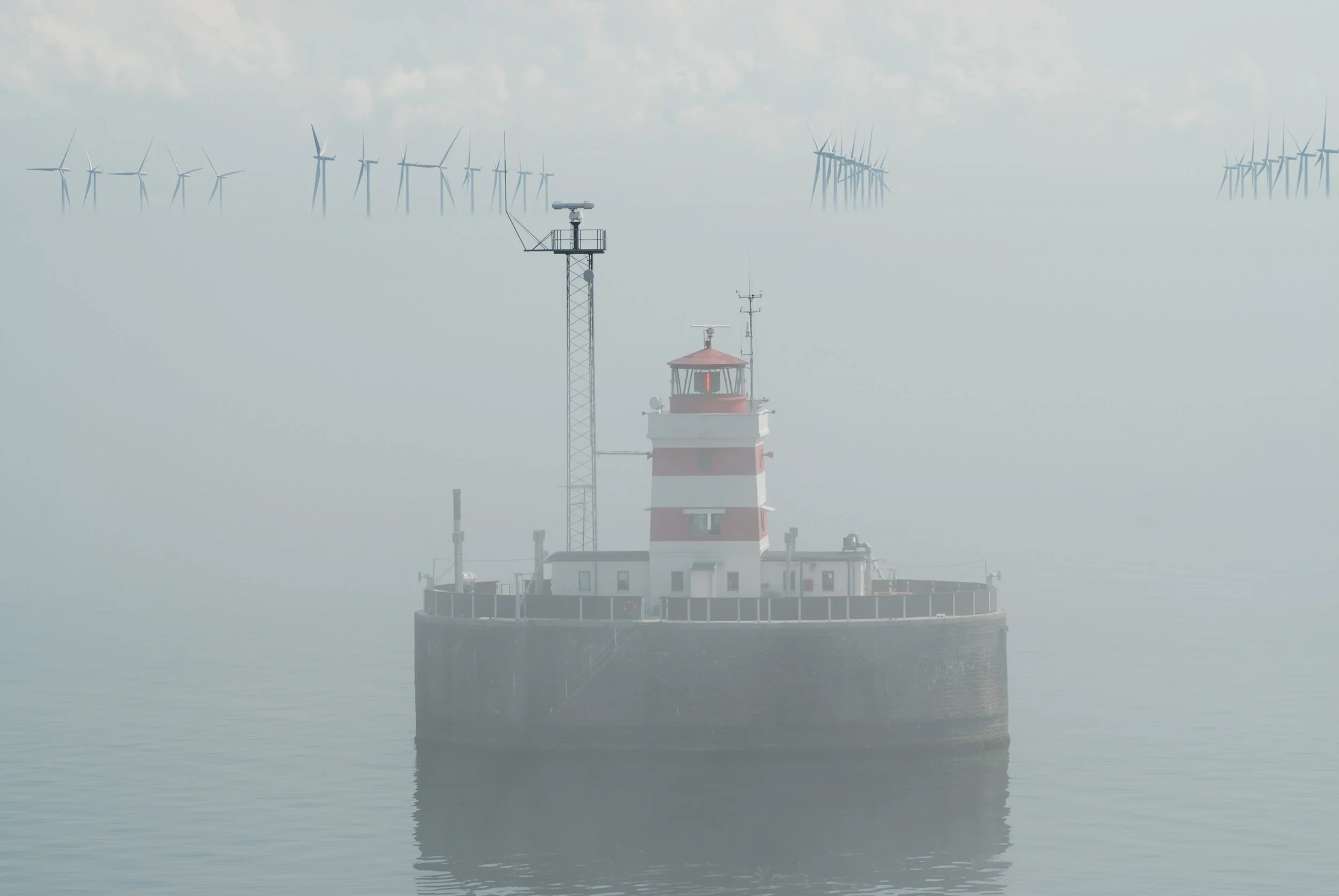 A lighthouse on a foggy body of water with wind turbines in the background.