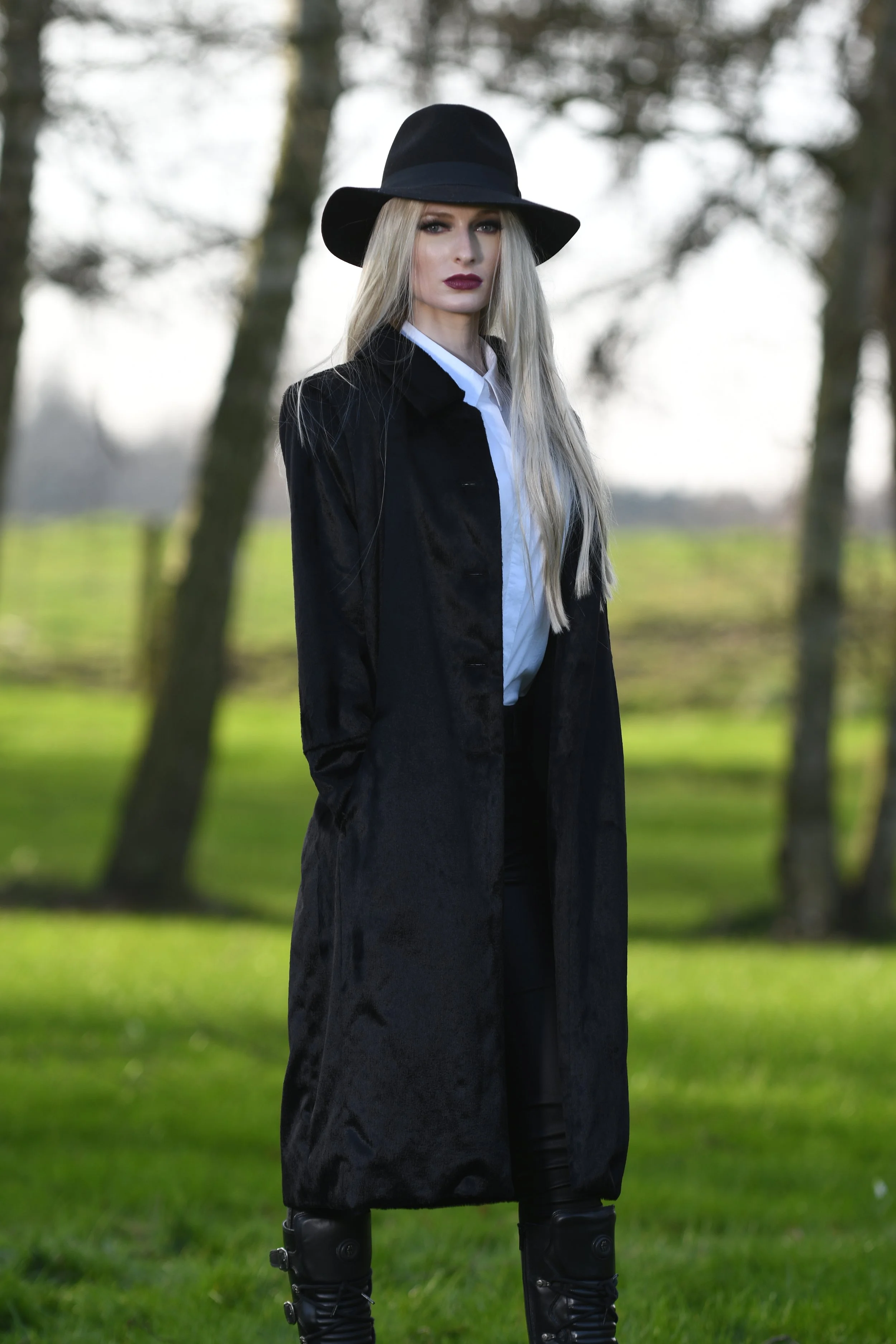 Woman with long blonde hair wearing a black hat, long black coat, white shirt, and black pants standing outdoors in a park with green grass and trees in the background.