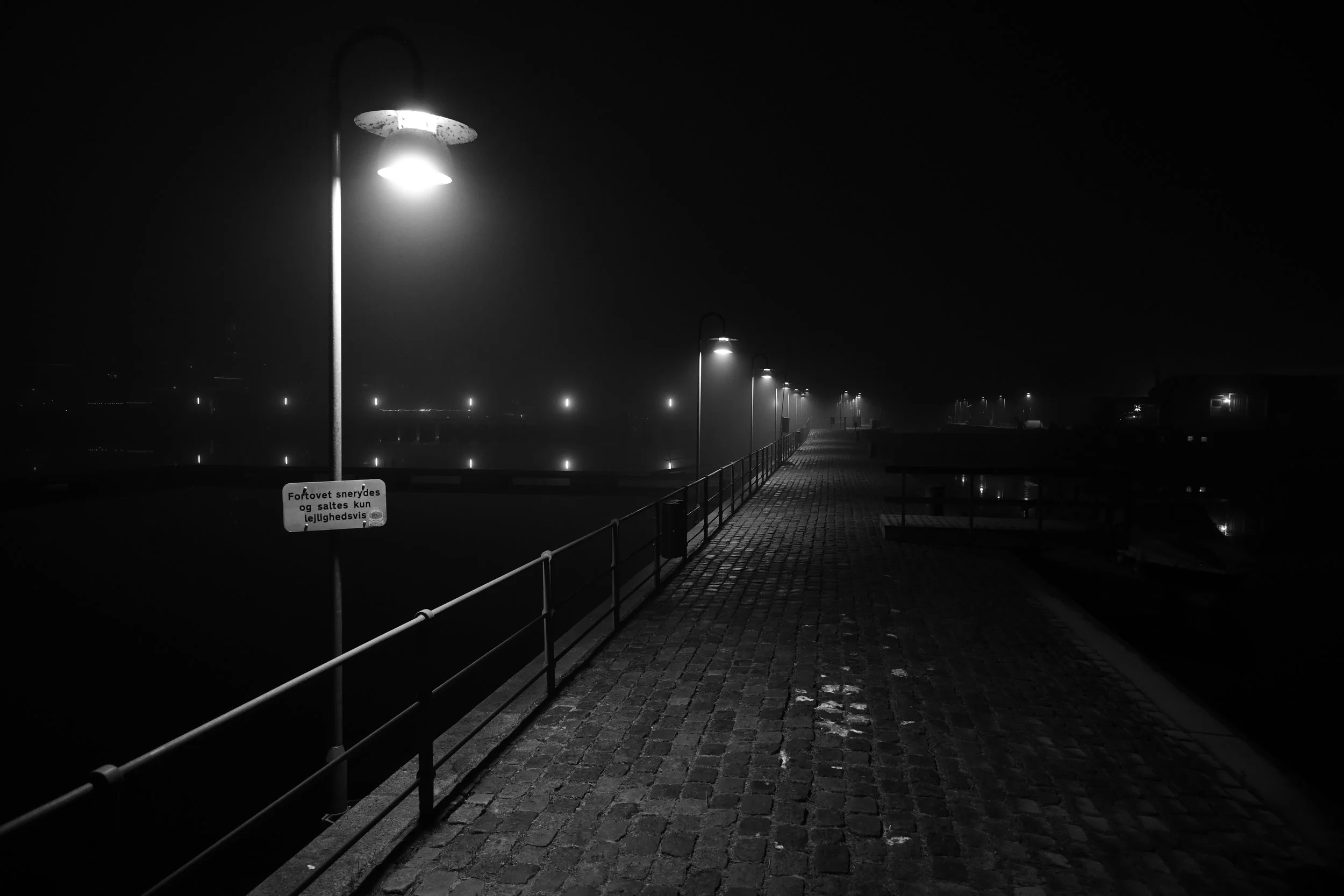 Empty cobblestone waterfront walkway illuminated by street lamps at night, with a foggy atmosphere and distant lights reflecting on the water.