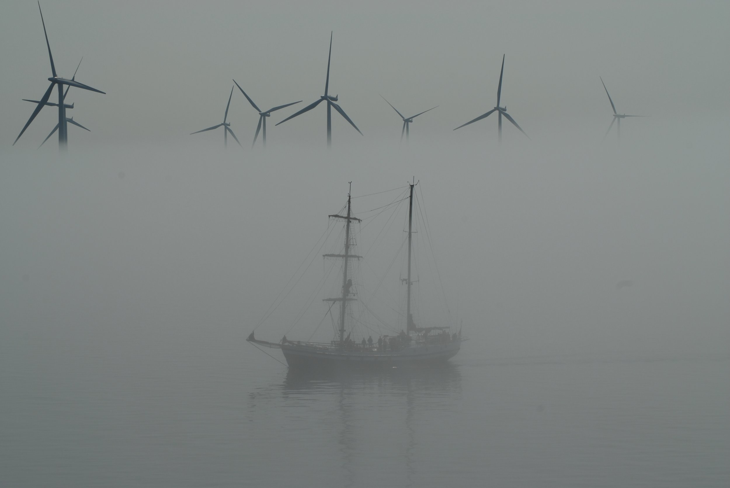 A sailing ship navigating through fog with a row of wind turbines in the background.