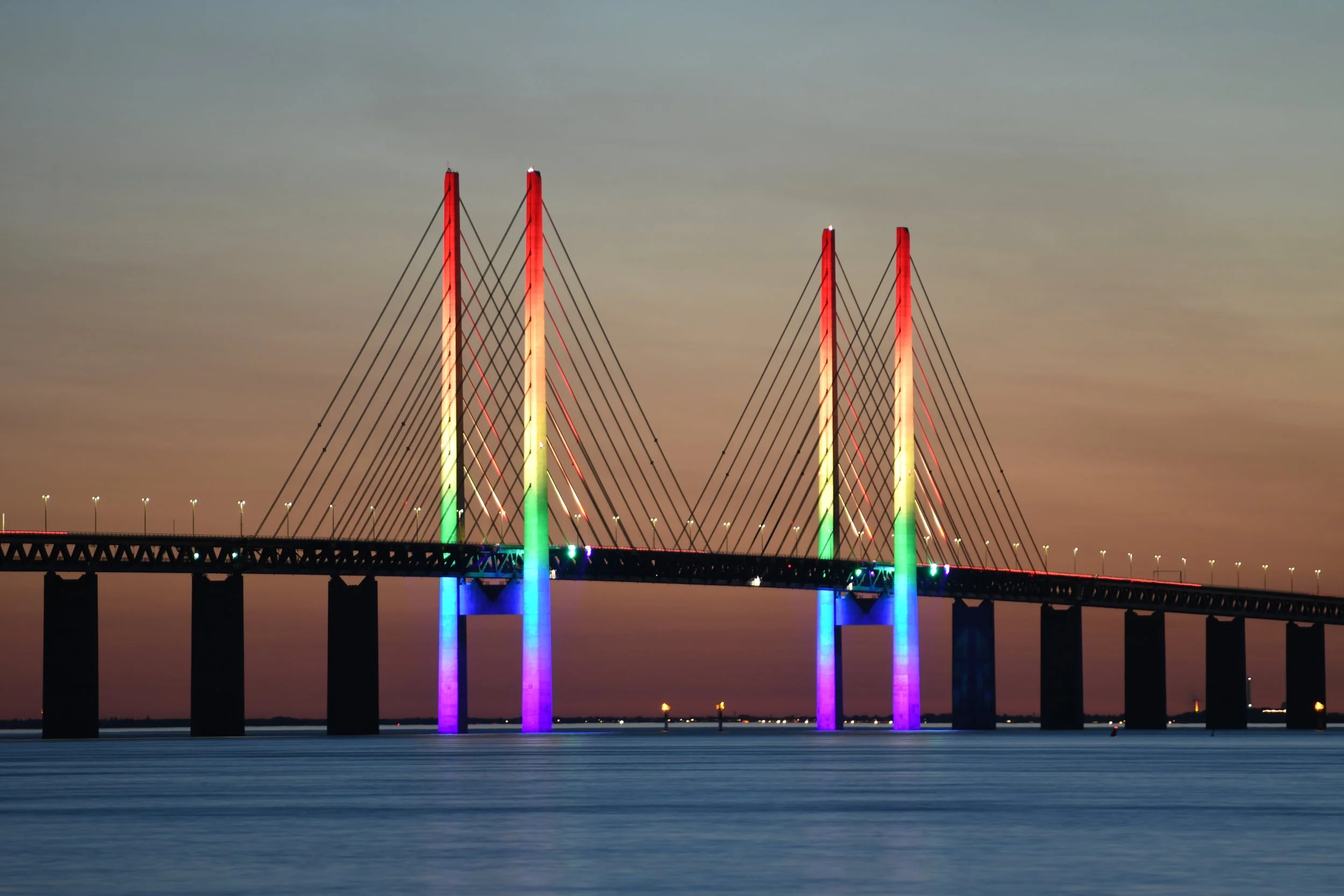 A bridge illuminated with multi-colored lights reflecting on the water at dusk.