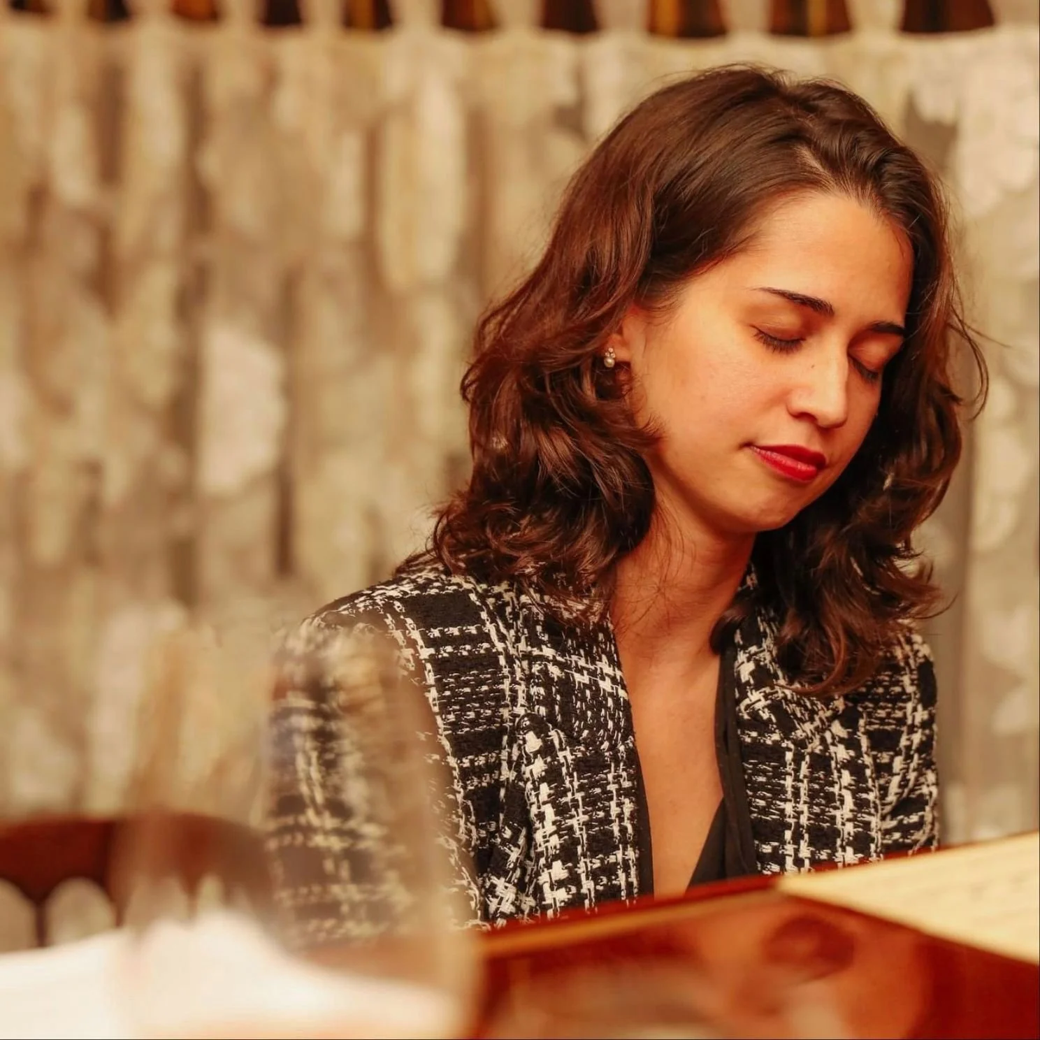 A woman with brown wavy hair and closed eyes sitting at a table, wearing a black and white patterned blazer, with a blurred glass and an open book or menu in foreground, against a beige curtain background.