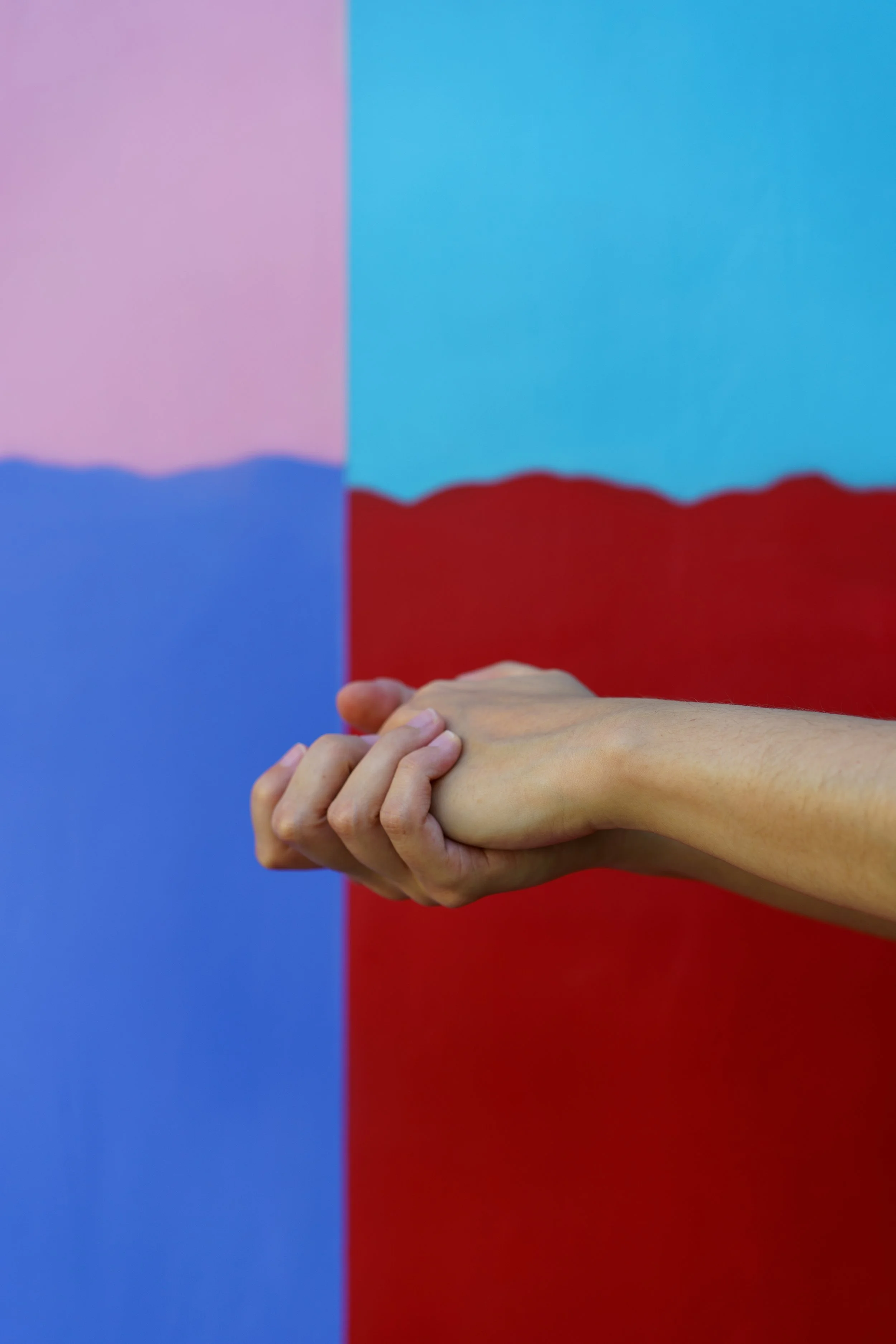 A person's arm and hand are holding onto another hand against a painted background with blue, red, and light pink sections.
