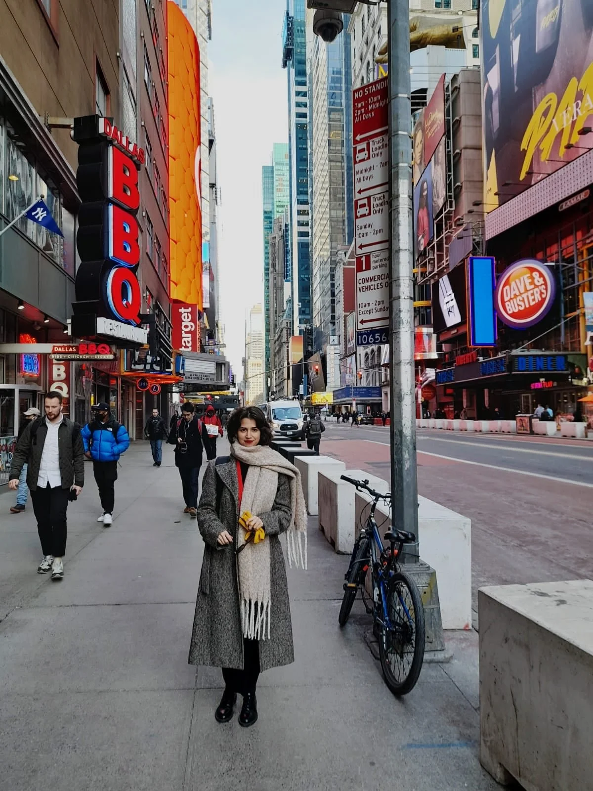 Woman standing on a city sidewalk in Times Square, New York City, wearing a long coat and scarf, with a bicycle parked beside her, surrounded by pedestrians and colorful neon signs and billboards.