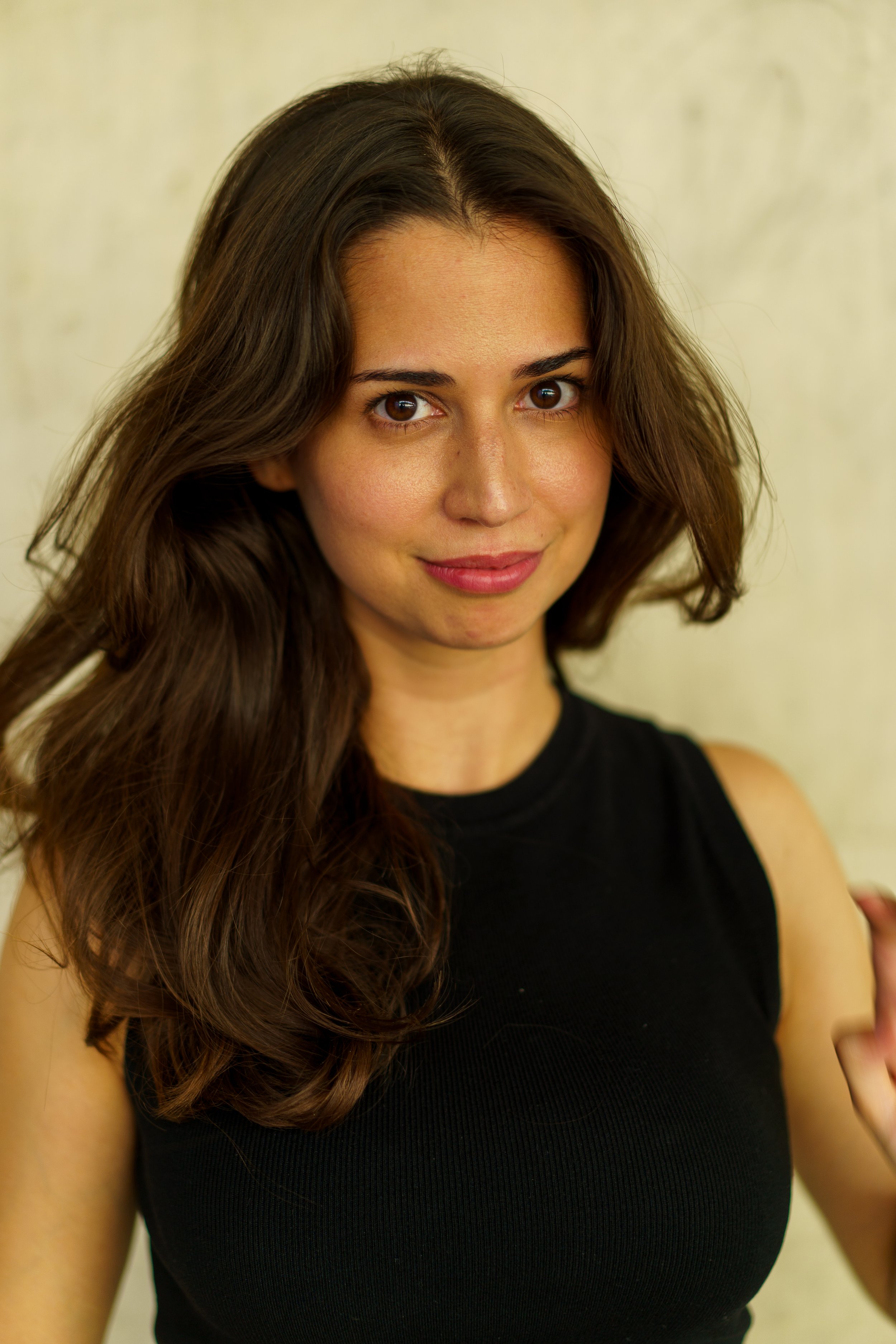 A young woman with long wavy brown hair, wearing a sleeveless black top, smiling gently at the camera against a plain light-colored background.