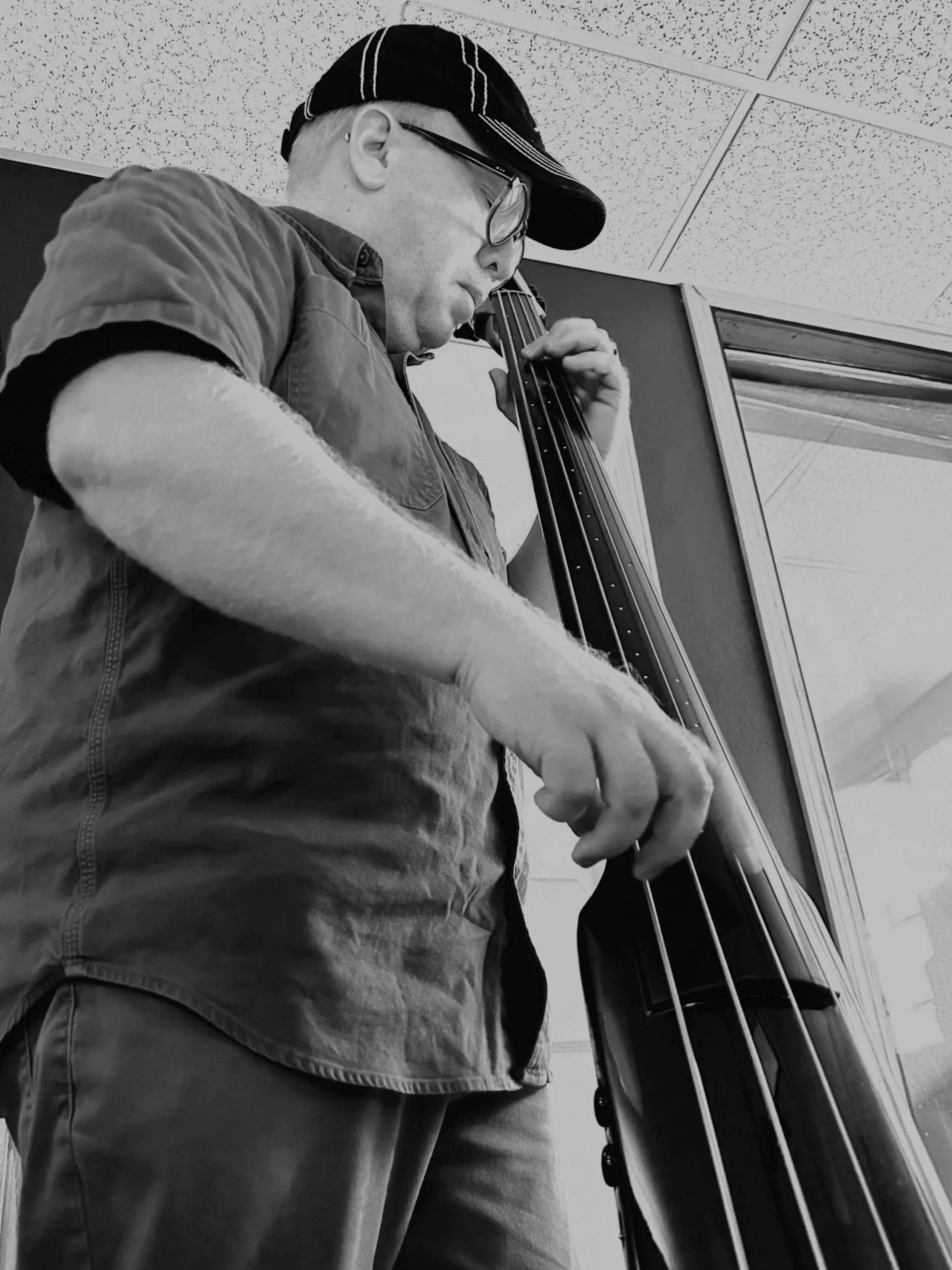 A man wearing glasses, a newsboy cap, and a short-sleeved shirt playing a double bass.