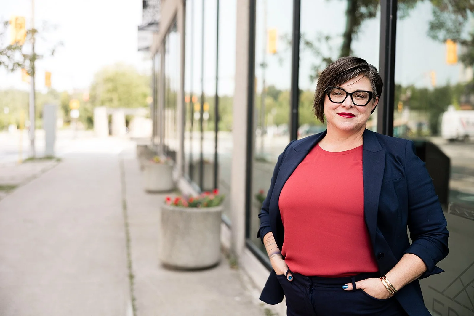 A woman with short dark hair, glasses, red lipstick, wearing a red shirt and a navy blazer, standing outdoors on a sidewalk with her hands in her pockets, smiling slightly, in front of a building with glass windows. There are trees and storefronts in the background.