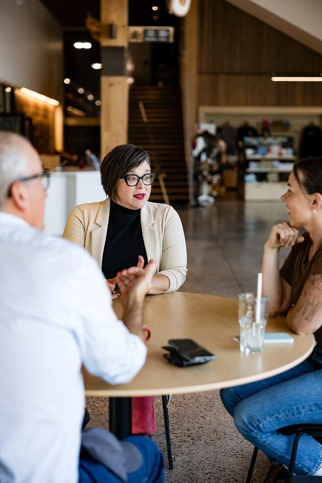 Three people sitting at a round table in a modern indoor space having a conversation. The woman in the center with short dark hair, glasses, and a beige blazer is speaking. The other man with gray hair and glasses, and the woman with a tattooed arm on the right are listening.