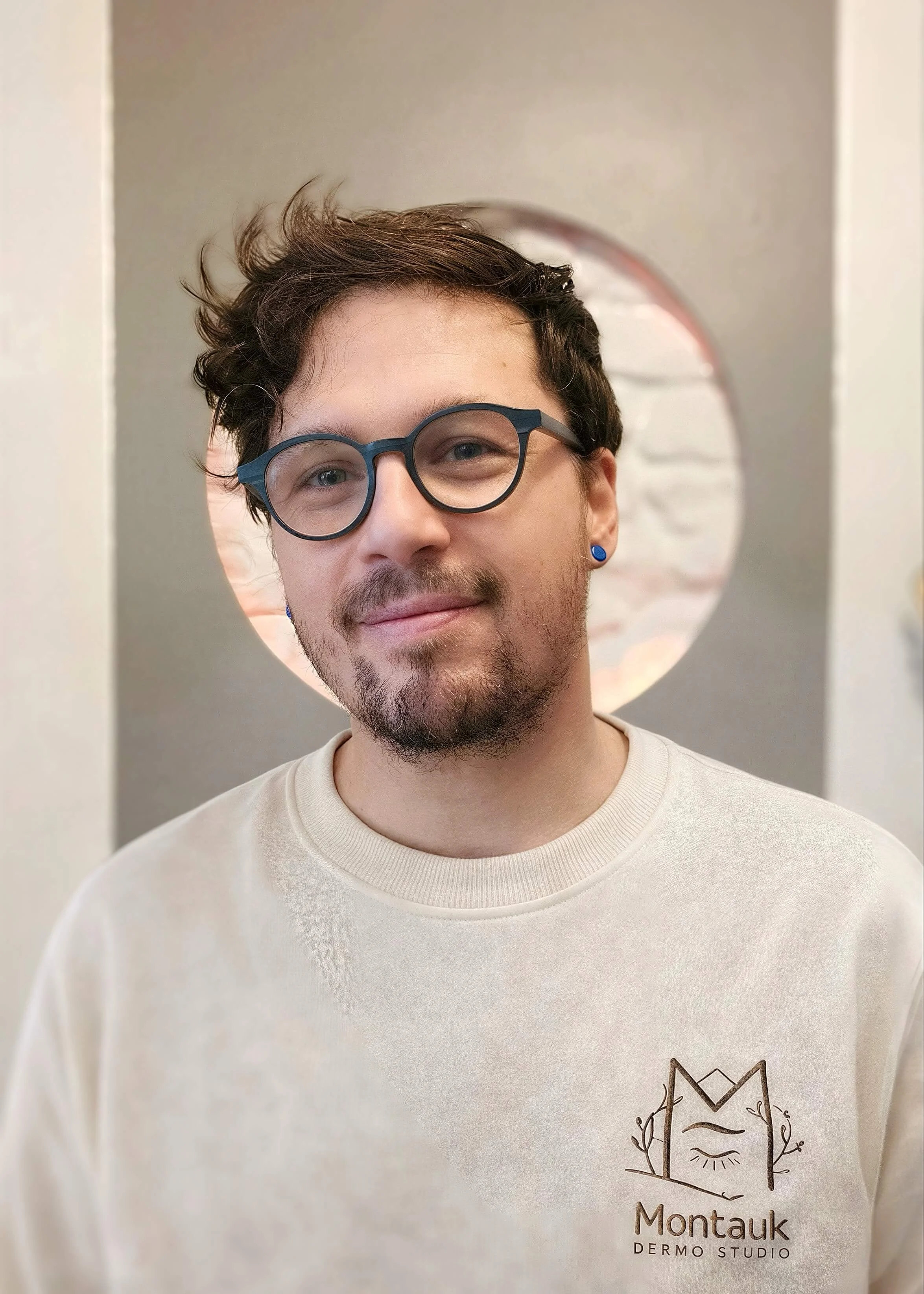 Portrait photo de Yohann, esthéticien et tatoueur à Nancy spécialisé dans la dermopigmentation et le maquillage semi permanent. Il porte des lunettes et un sweat shirt affichant le logo "Montauk dermo studio". La photo est douce et professionnelle.