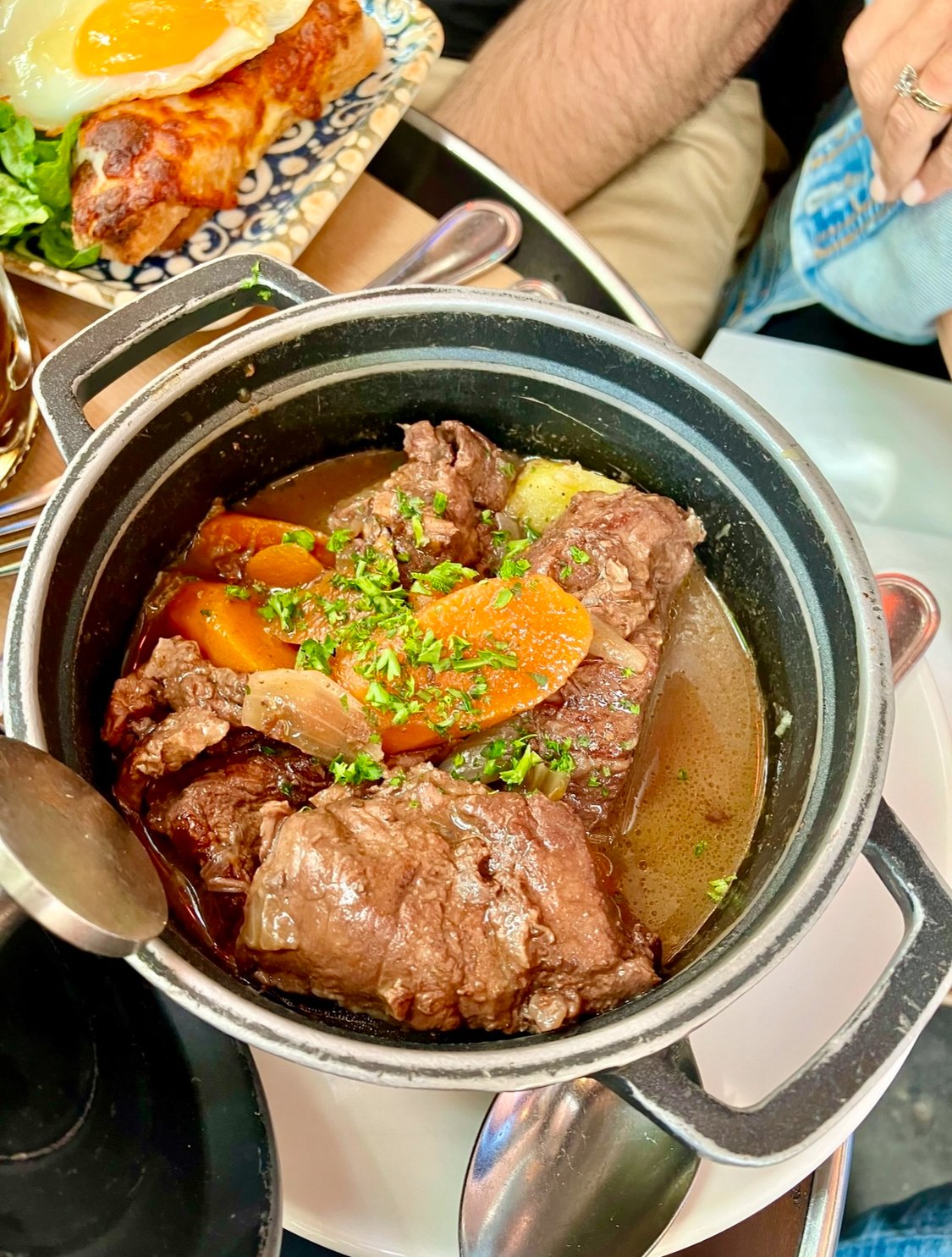 A pot of beef stew with carrots, potatoes, and chopped parsley, with a plate of breakfast meat topped with a fried egg and greens in the background.