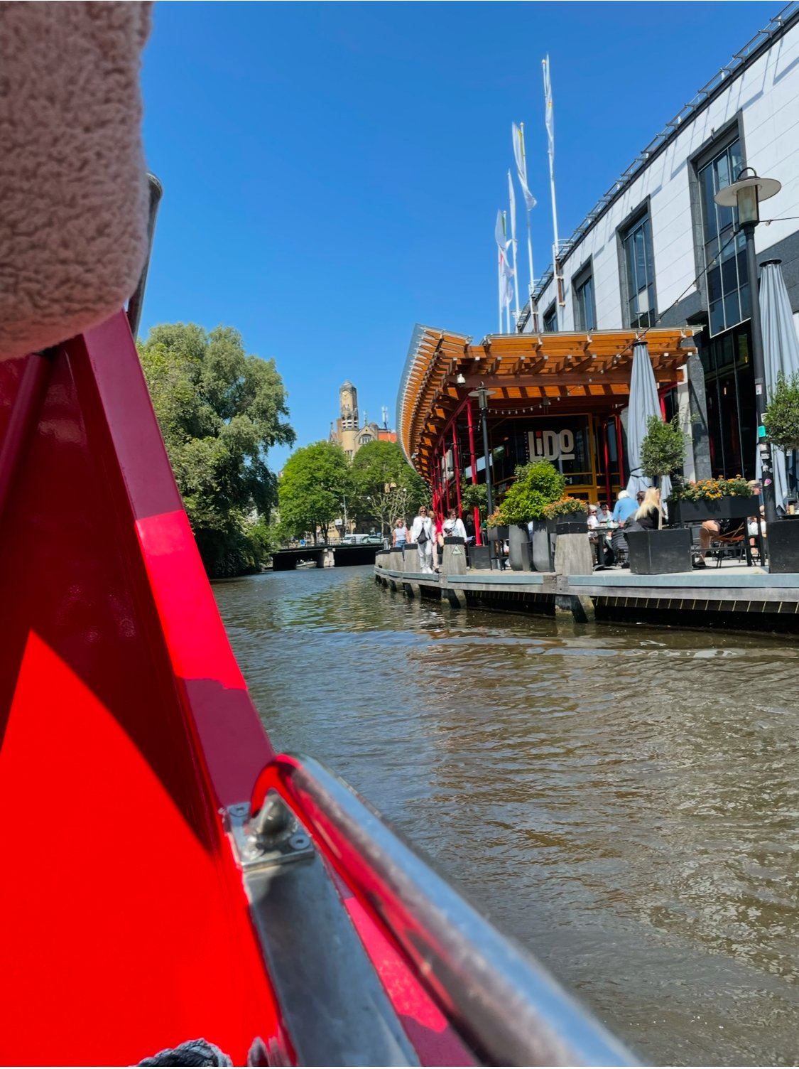 A view from a boat on a river showing a lively riverside cafe with outdoor seating, modern architecture, flags, trees, and a historic building in the background under a clear blue sky.