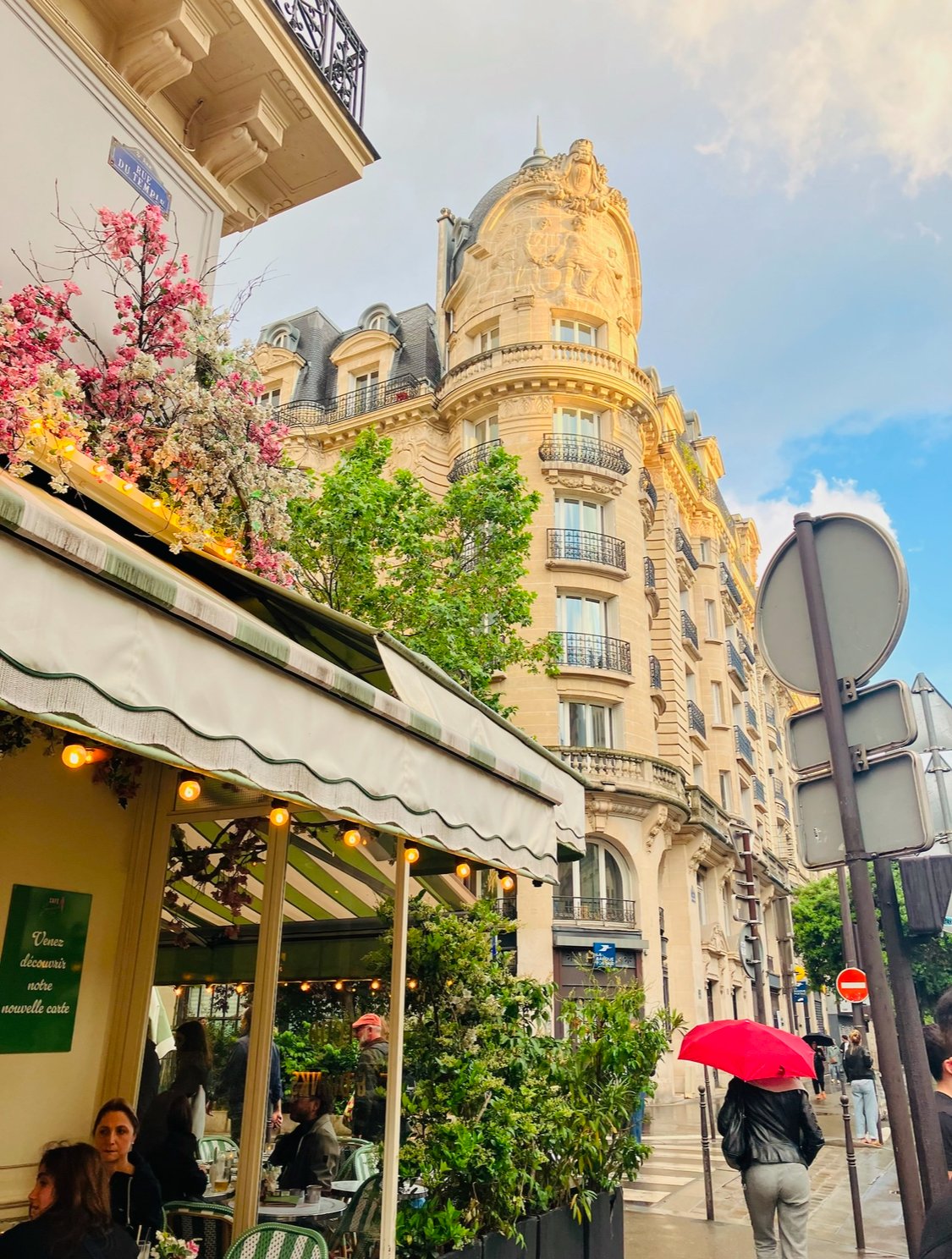 Parisian street scene with a restaurant outdoor seating area, a pink umbrellas, classic Haussmann-style buildings, and blooming trees under a partly cloudy sky.