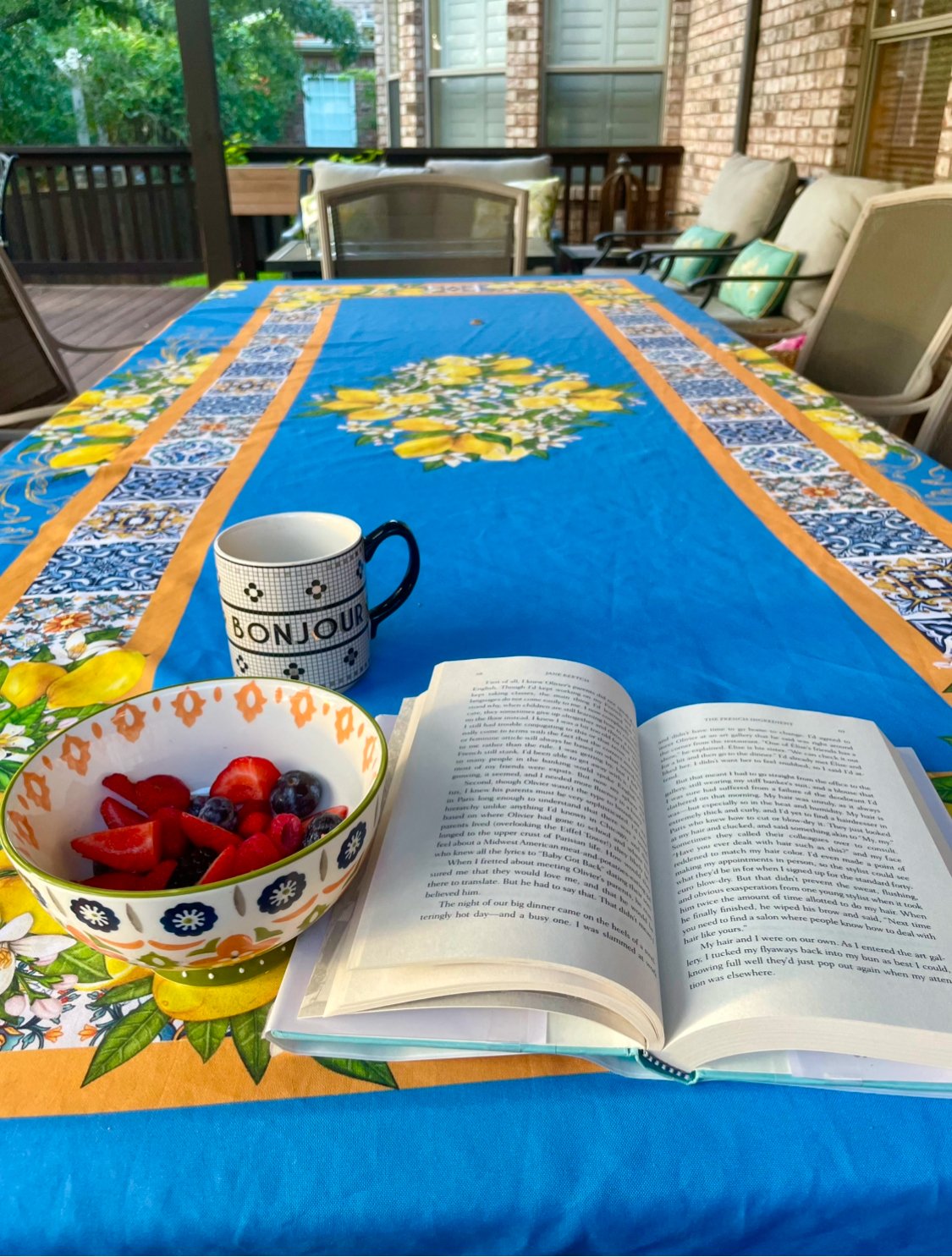 An outdoor dining table with a colorful floral tablecloth, a bowl of mixed berries, a coffee mug labeled 'Bonjour', and an open book, set on a screened porch with chairs and a view of trees outside.
