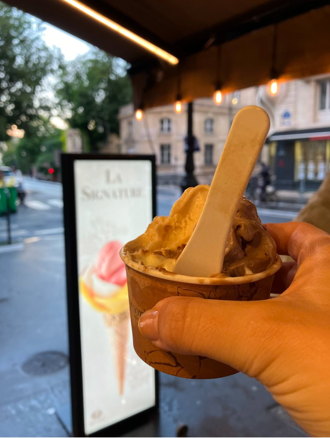 Hand holding a cup of ice cream with a white spoon, outside near a sign and building in the background during dusk.