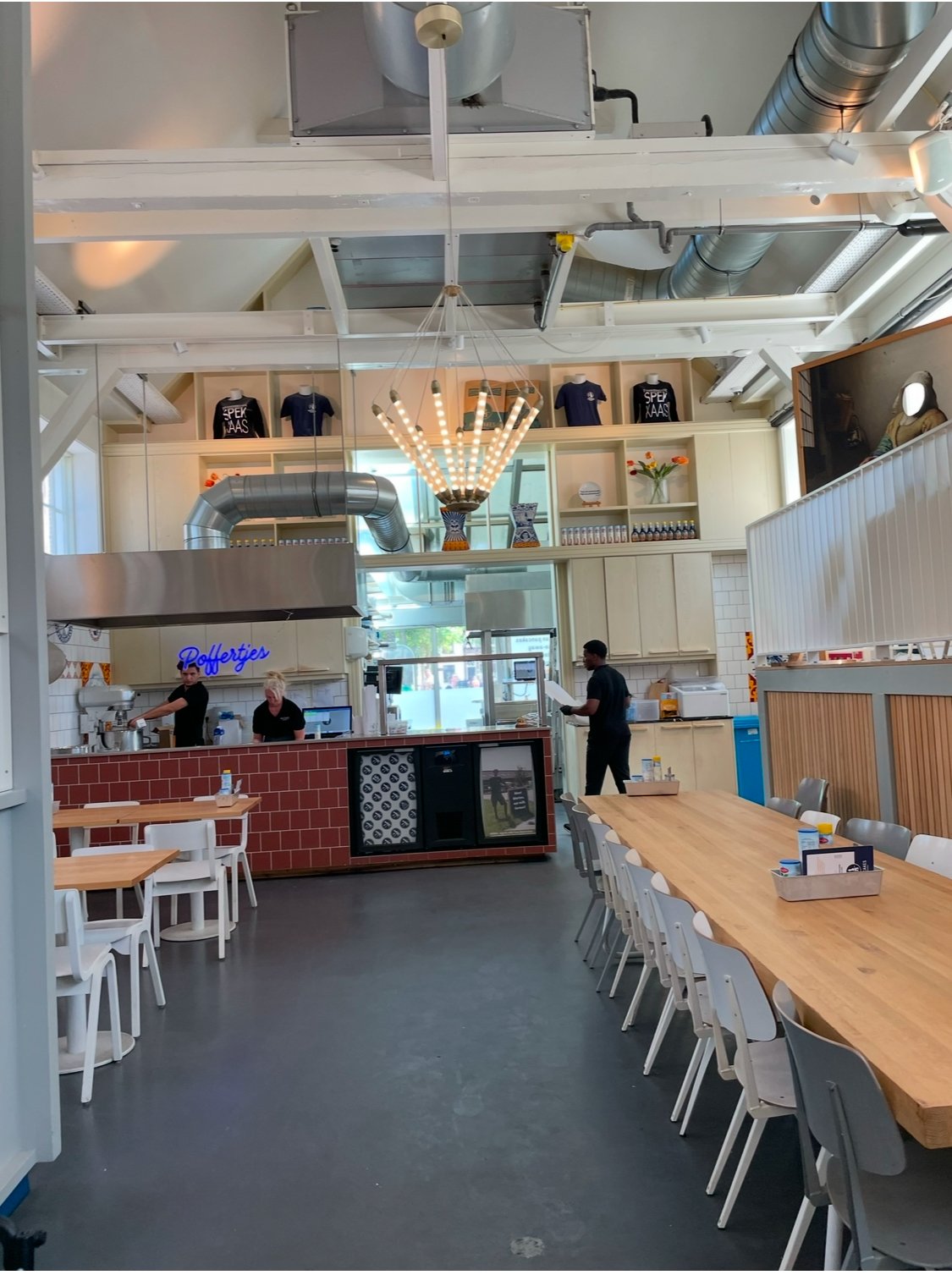 Interior of a modern, casual restaurant with wooden tables and white chairs, a counter with staff, and decorative elements like a chandelier and shelves with shirts, vases, and bottles.