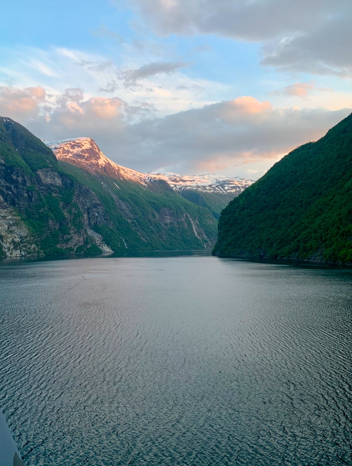 A picturesque fjord surrounded by steep, green mountains with snow-capped peaks in the background, under a partly cloudy sky.