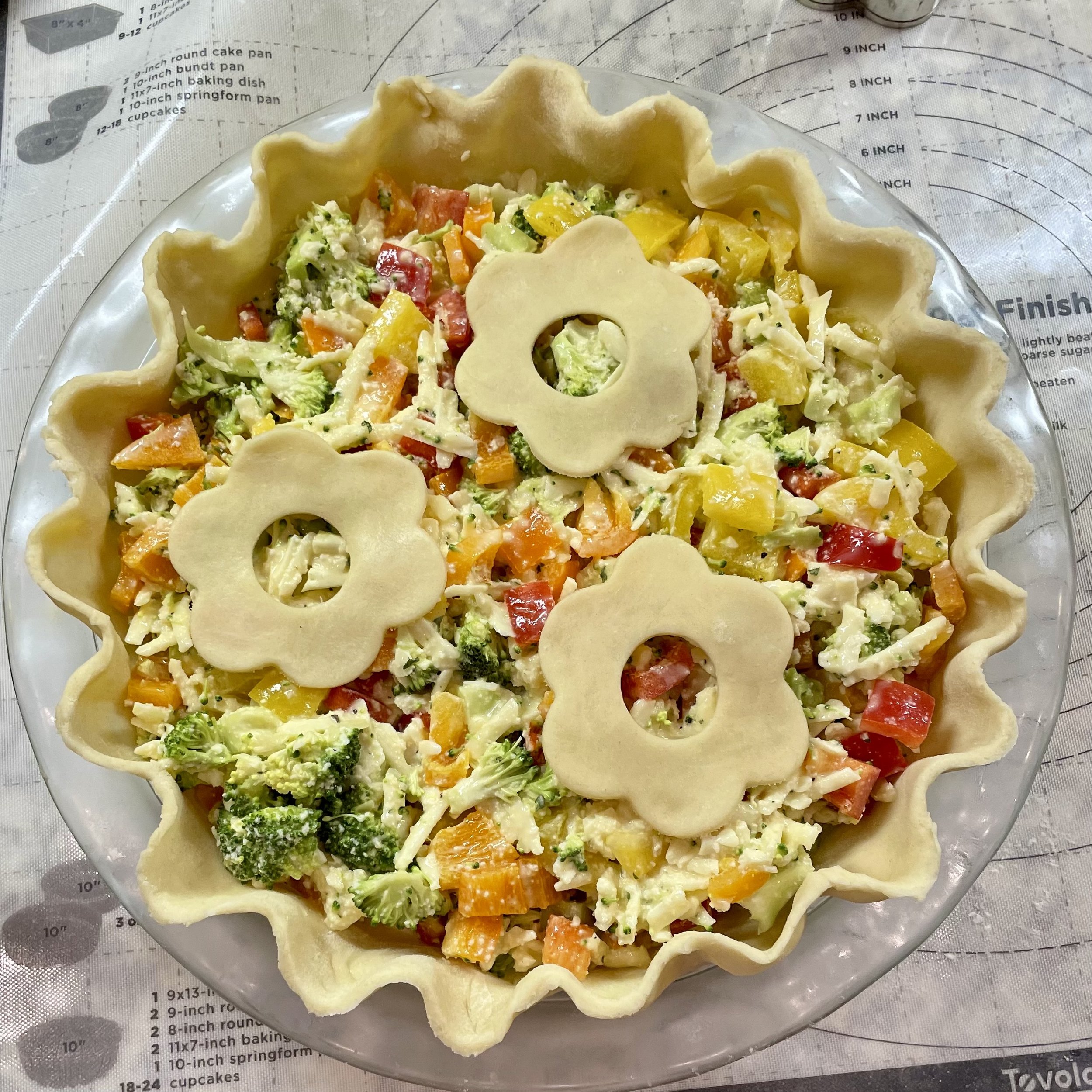 A partially assembled vegetable quiche with a decorative pastry crust, filled with chopped broccoli, red and yellow bell peppers, shredded cheese, and topped with three flower-shaped pastry cutouts.