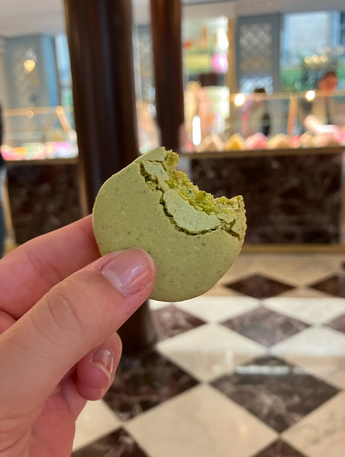 Close-up of a hand holding a cracked green macaron inside a bakery or cafe with customers and display cases in the background.
