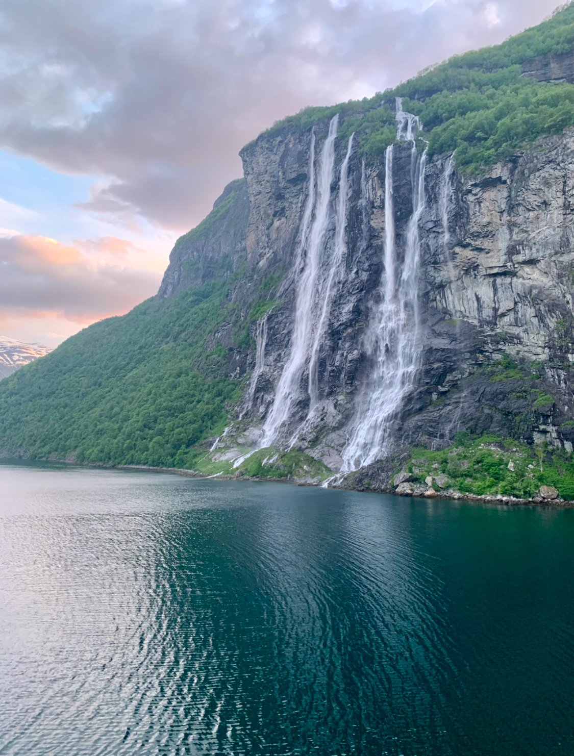 Waterfalls cascading down a rocky cliff next to a calm body of water with green hills and a cloudy sky in the background.