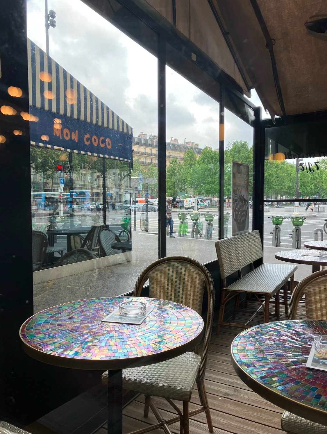 Interior of a café with colorful mosaic tables, chairs, and windows looking out onto a city street with trees, buildings, and bicycles.