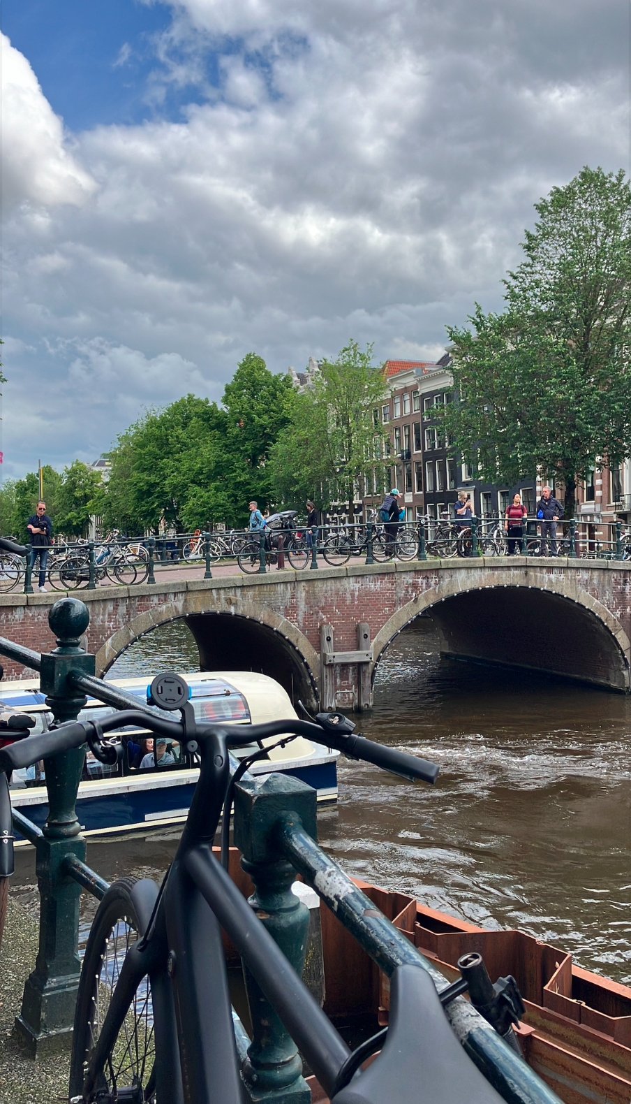 A scene of a canal in Amsterdam with a bridge, bicycles, and boats, under a cloudy sky.