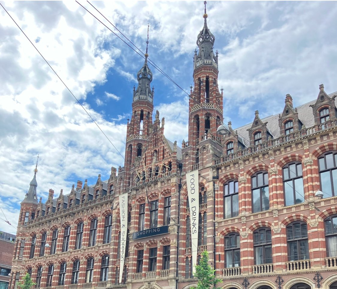 A historic Gothic-style building with red and cream striped brickwork, tall spires, and arched windows under a partly cloudy sky, with banners advertising shopping, food, and drinks.