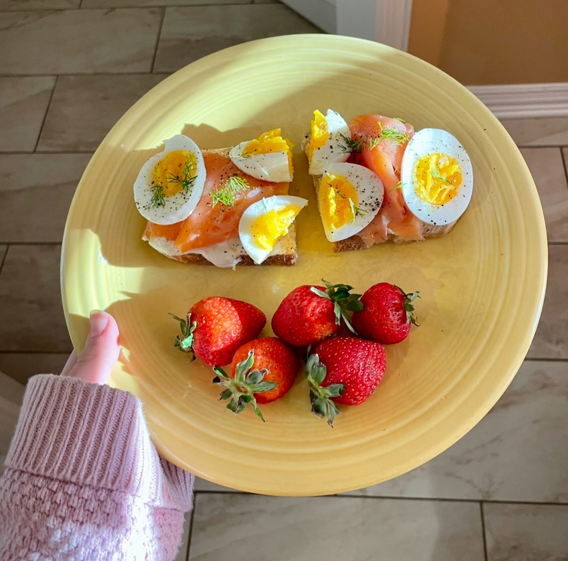 A yellow plate with two open-faced eggs Benedict with smoked salmon and dill, and a bunch of fresh strawberries. A person's hand in a pink sweater is holding the plate.