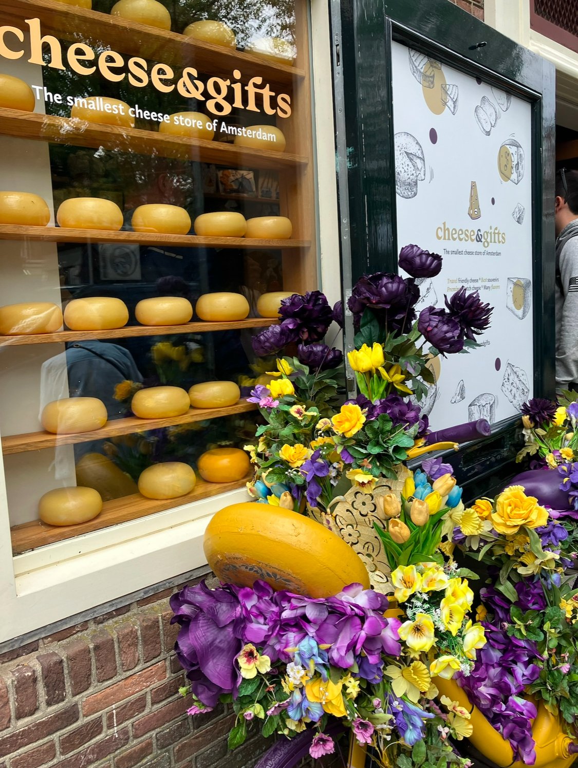 Display of various cheeses in a shop window with a sign reading 'cheese & gifts, The smallest cheese store of Amsterdam,' decorated with colorful flowers.