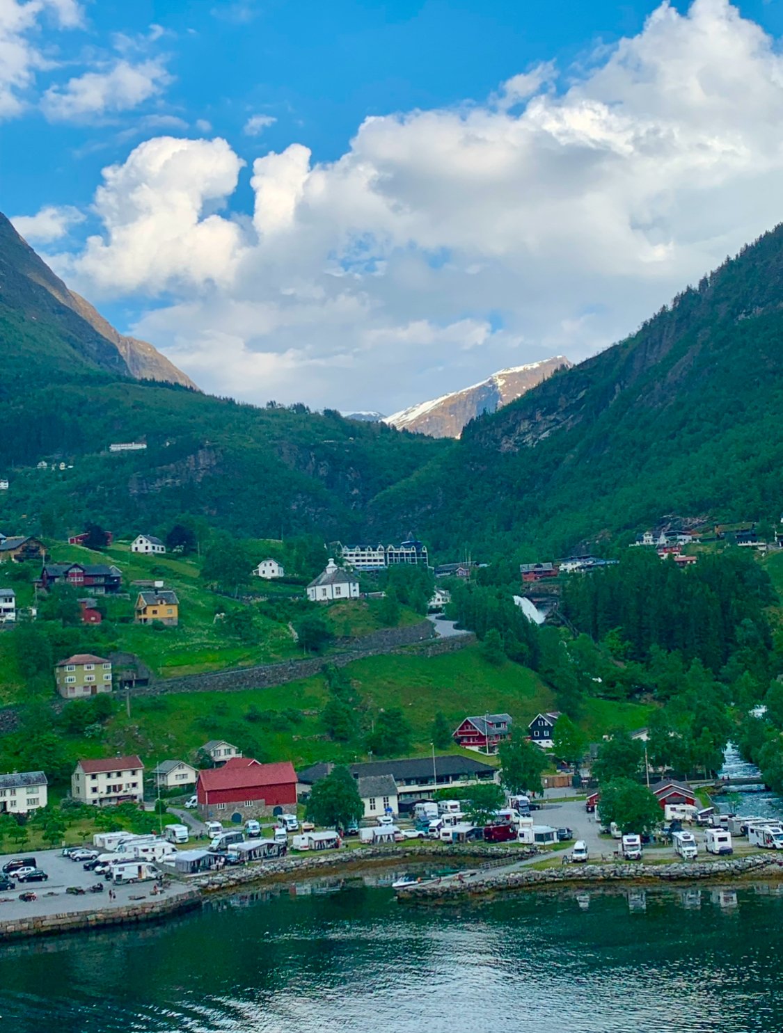 Scenic view of a small coastal village nestled in green mountains, with colorful houses, a marina with boats, and a waterfall in the background under a partly cloudy sky.