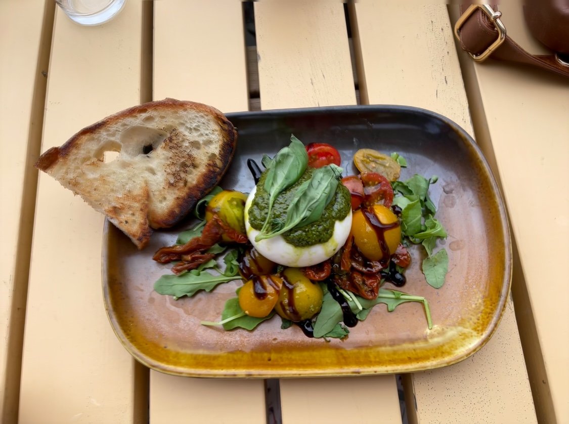 Caprese salad with cherry tomatoes, fresh basil, mozzarella, balsamic glaze, and a side slice of toasted bread on a brown ceramic plate.