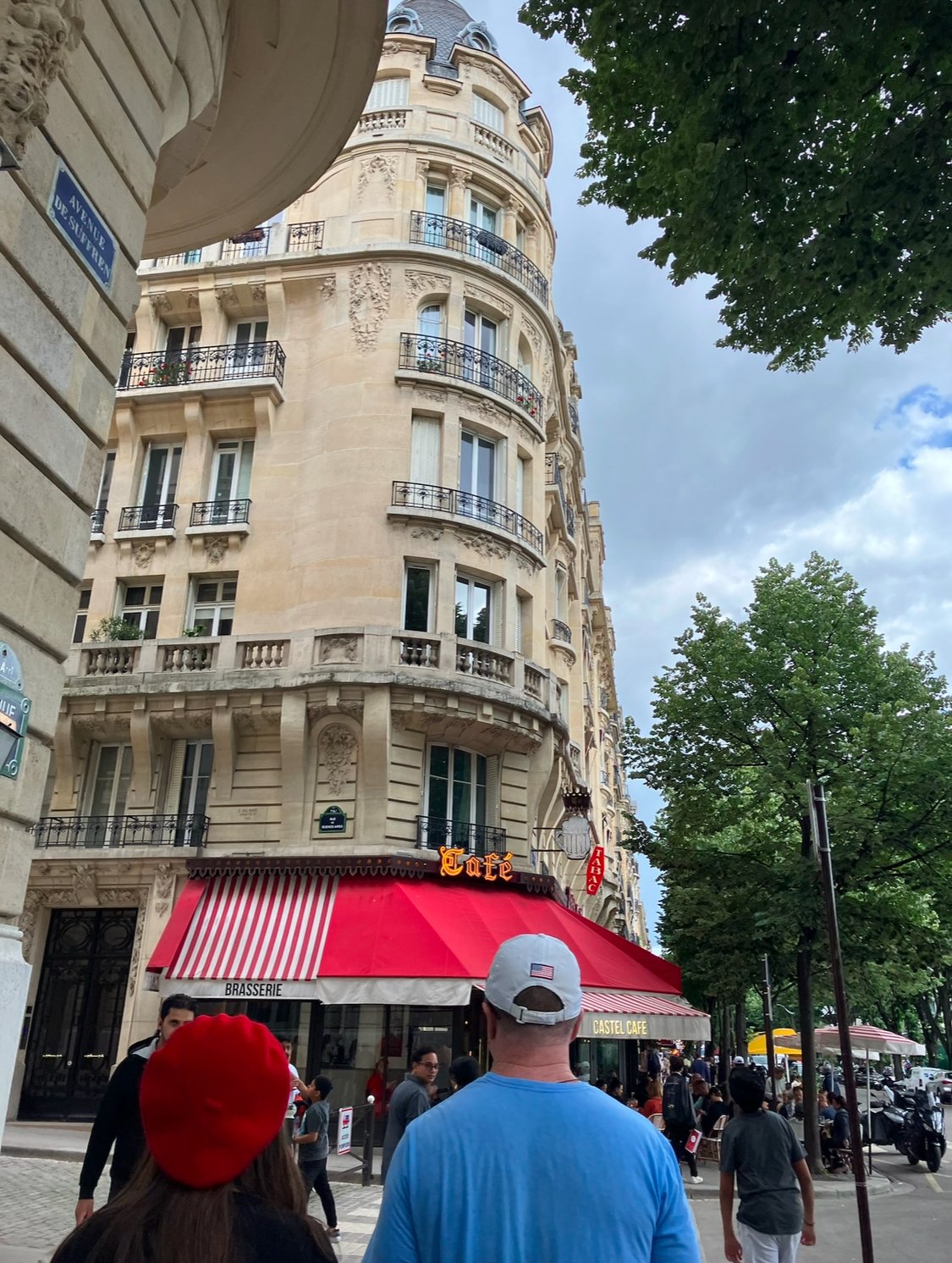 People walking past a cafe called Castal in a Parisian street, with classic Paris apartment buildings and trees lining the sidewalk.