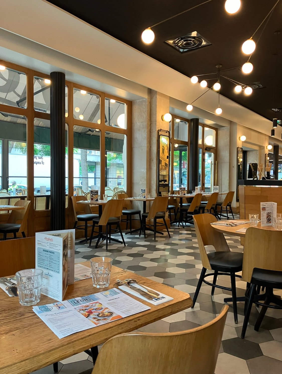 Empty modern restaurant with wooden tables and chairs, large windows, and hexagonal floor tiles, decorated with warm lighting.