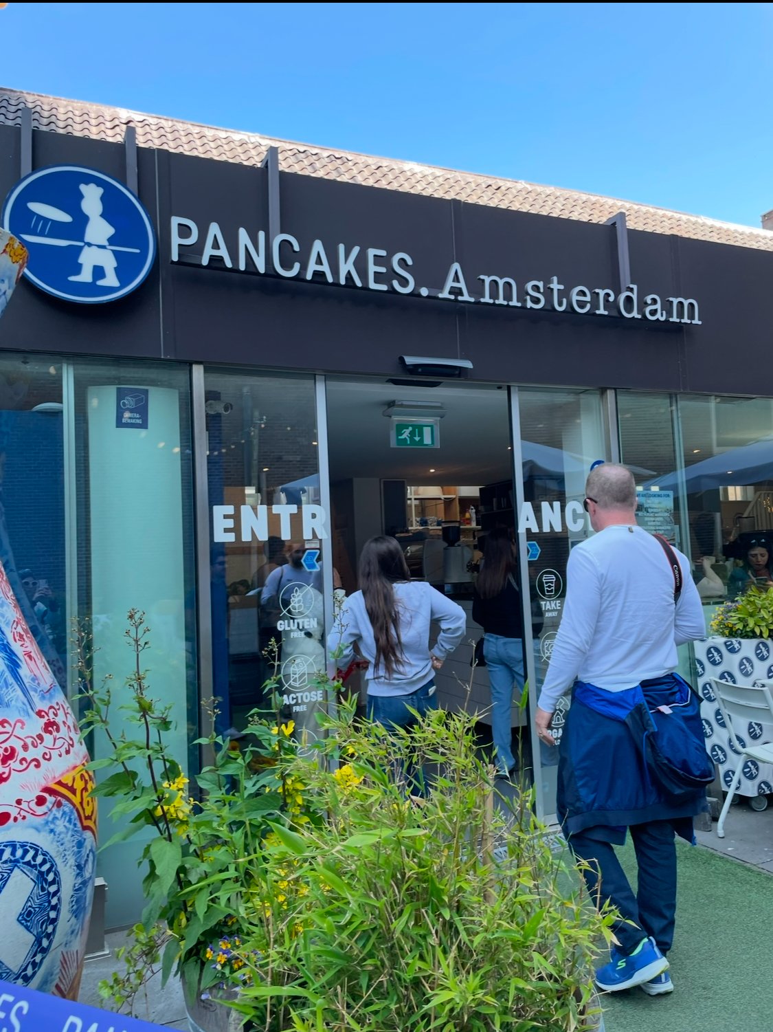 People standing outside a pancake restaurant named Pancakes Amsterdam with glass doors, visible signage for gluten-free and lactose-free options, and a greenery plant in the foreground.
