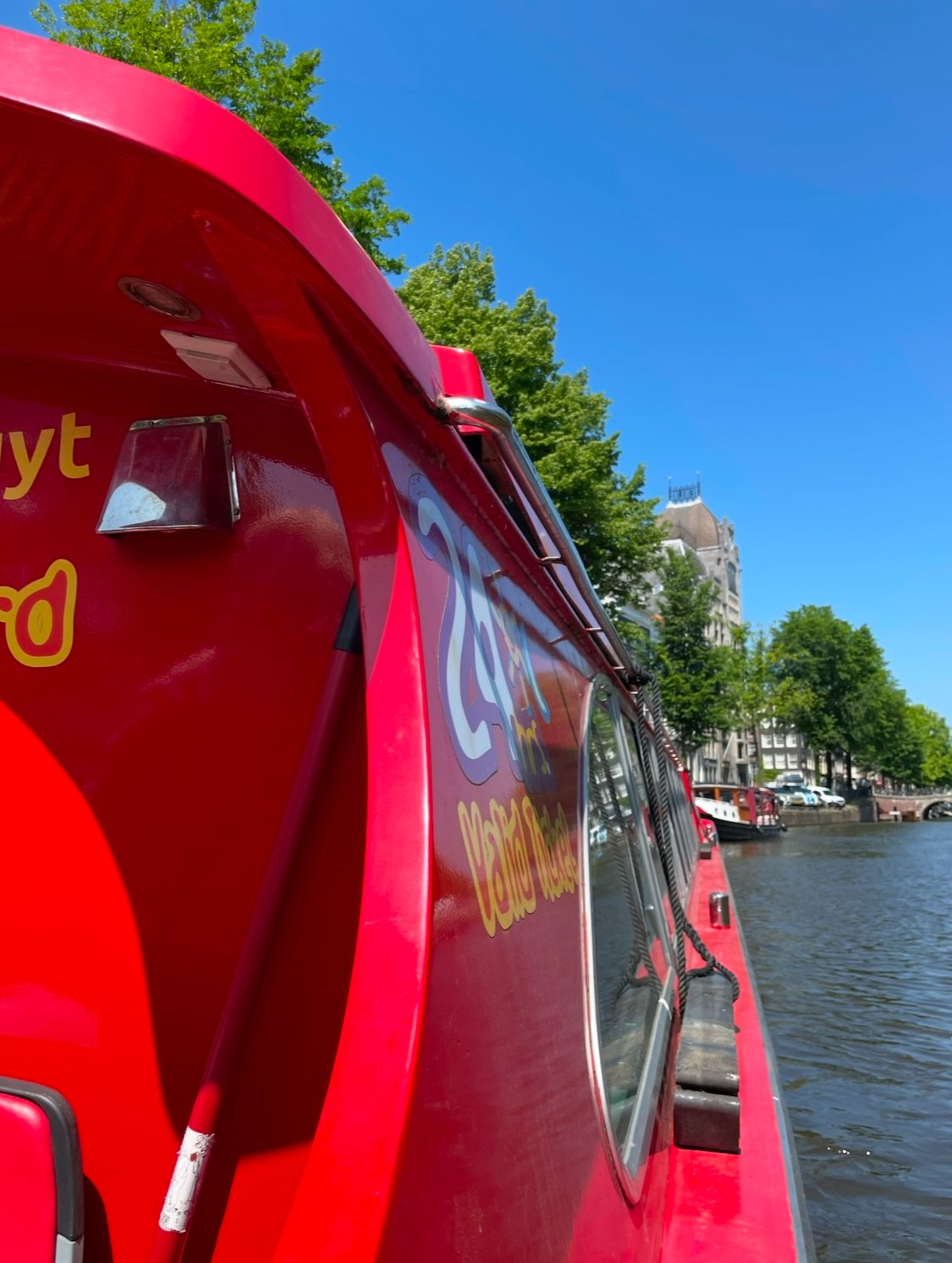 Red boat on a river with green trees and buildings in the background under a clear blue sky.