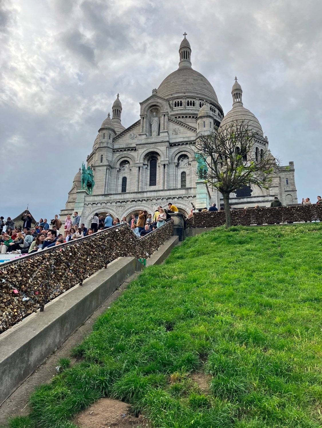 People gathered outside the Sacré-Cœur Basilica on a cloudy day in Montmartre, Paris, France, with a grassy area, a tree, and a sky filled with clouds.