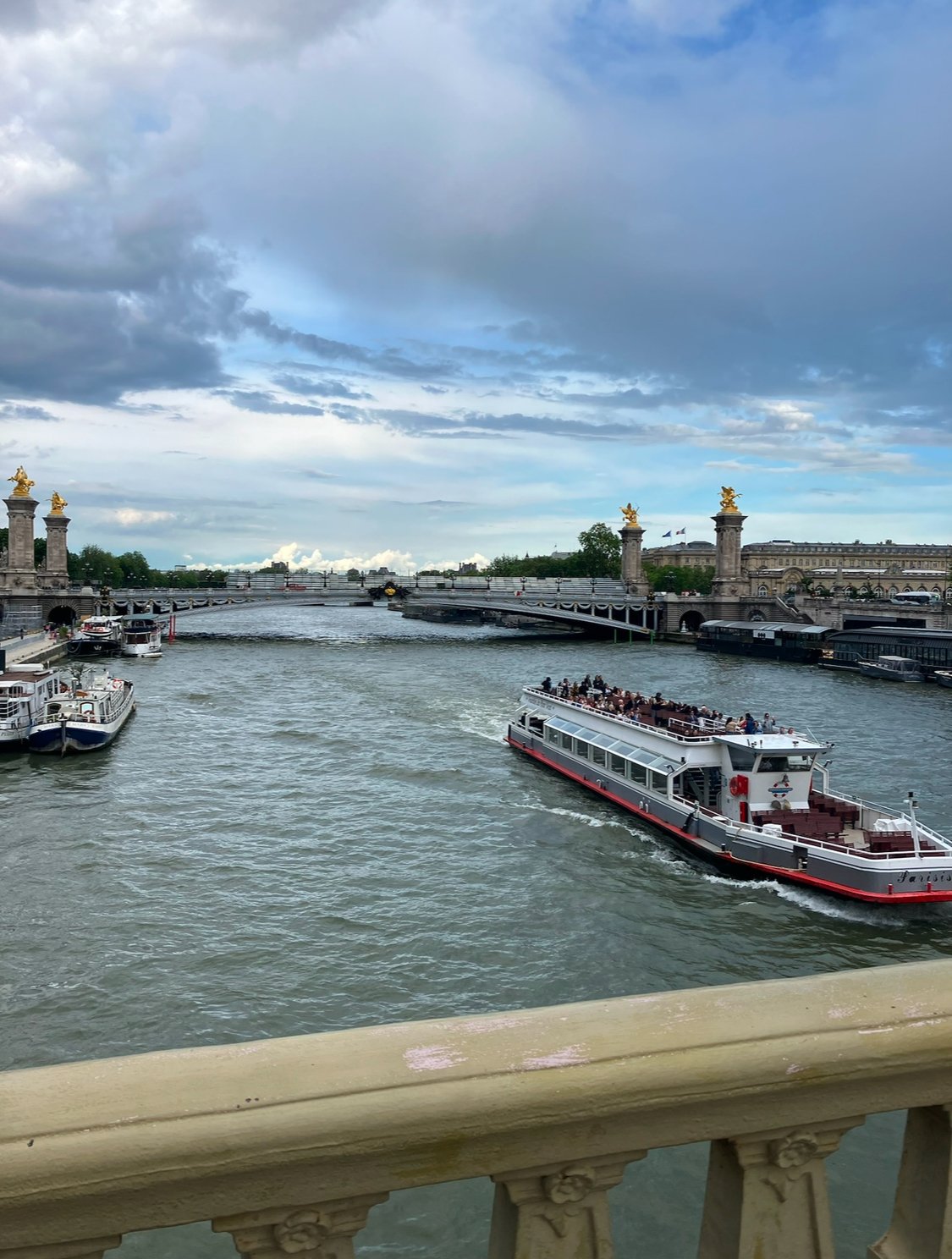 A river scene with boats, including a large sightseeing boat, passing under a bridge with golden statues on top, during daytime with cloudy sky.