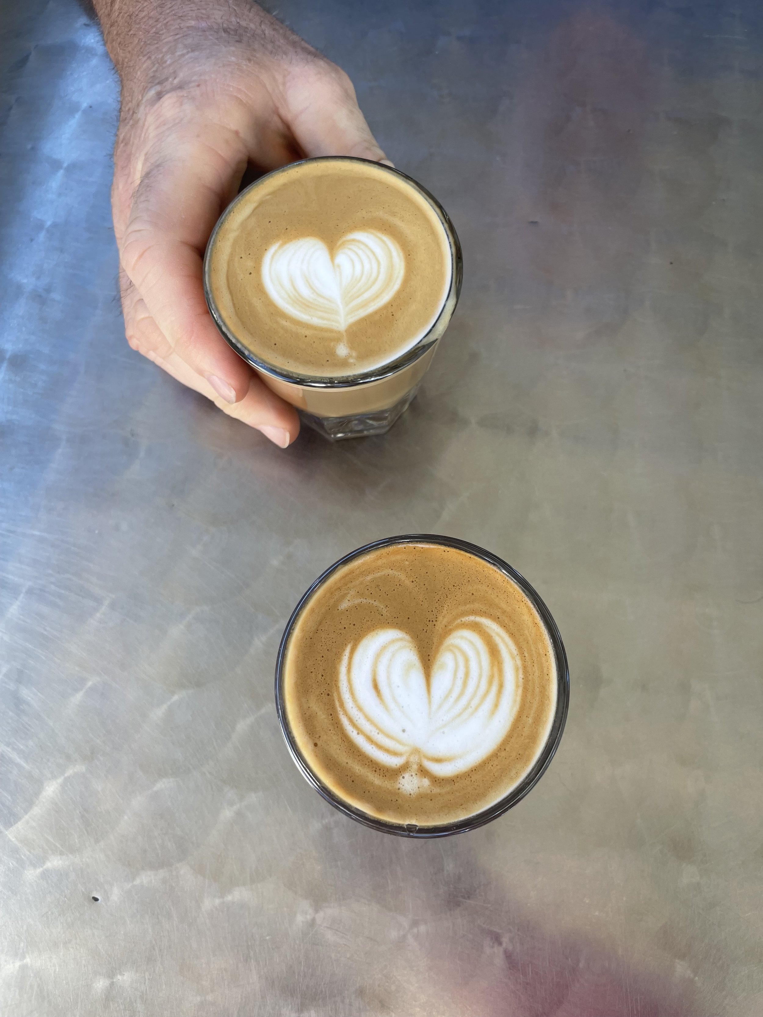 Two glasses of latte with heart-shaped latte art on a metal table, one being held by a hand.