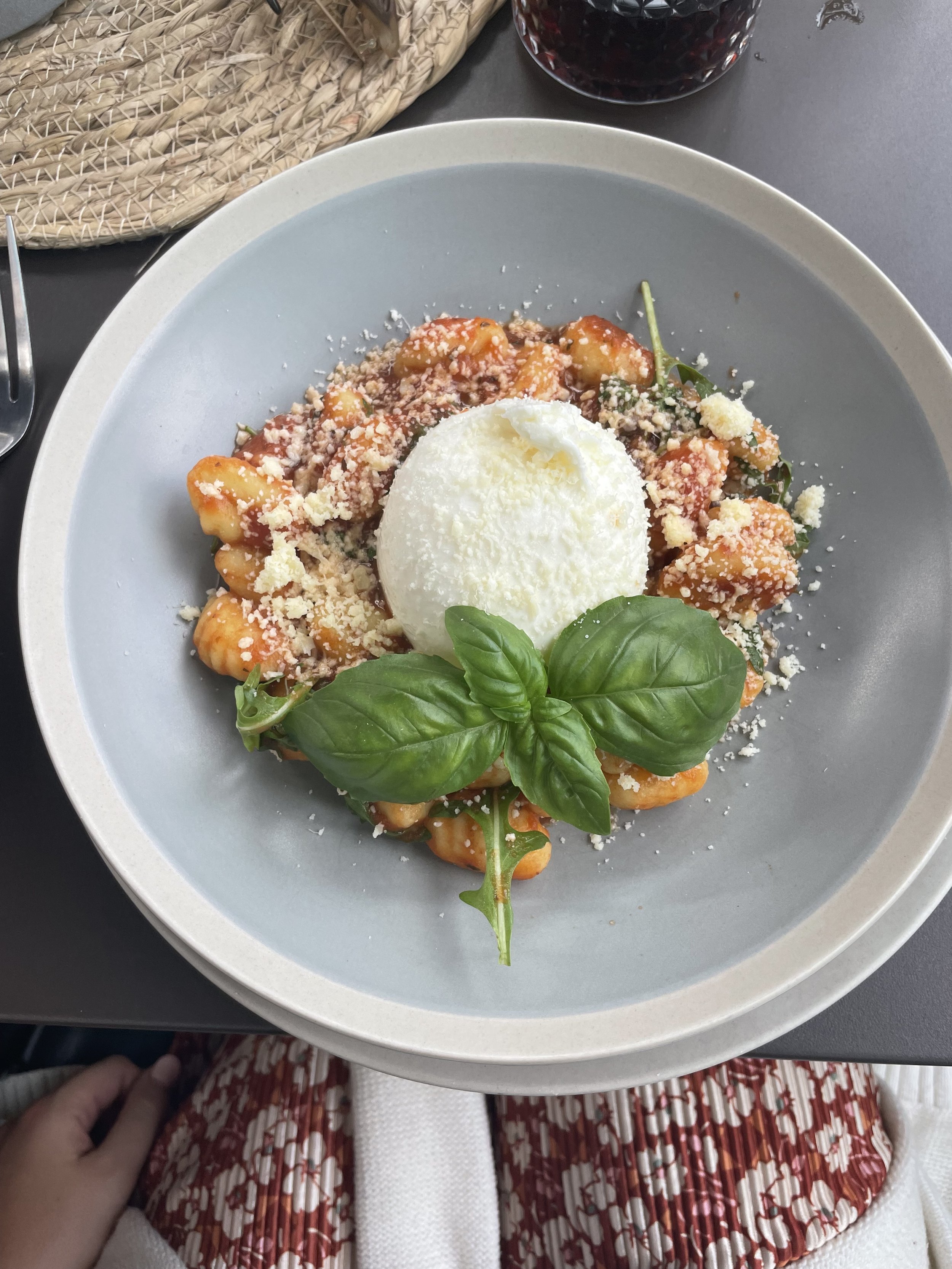 Plate of gnocchi pasta topped with tomato sauce, grated cheese, a scoop of ricotta cheese, and fresh basil leaves, with a glass of red wine in the background.
