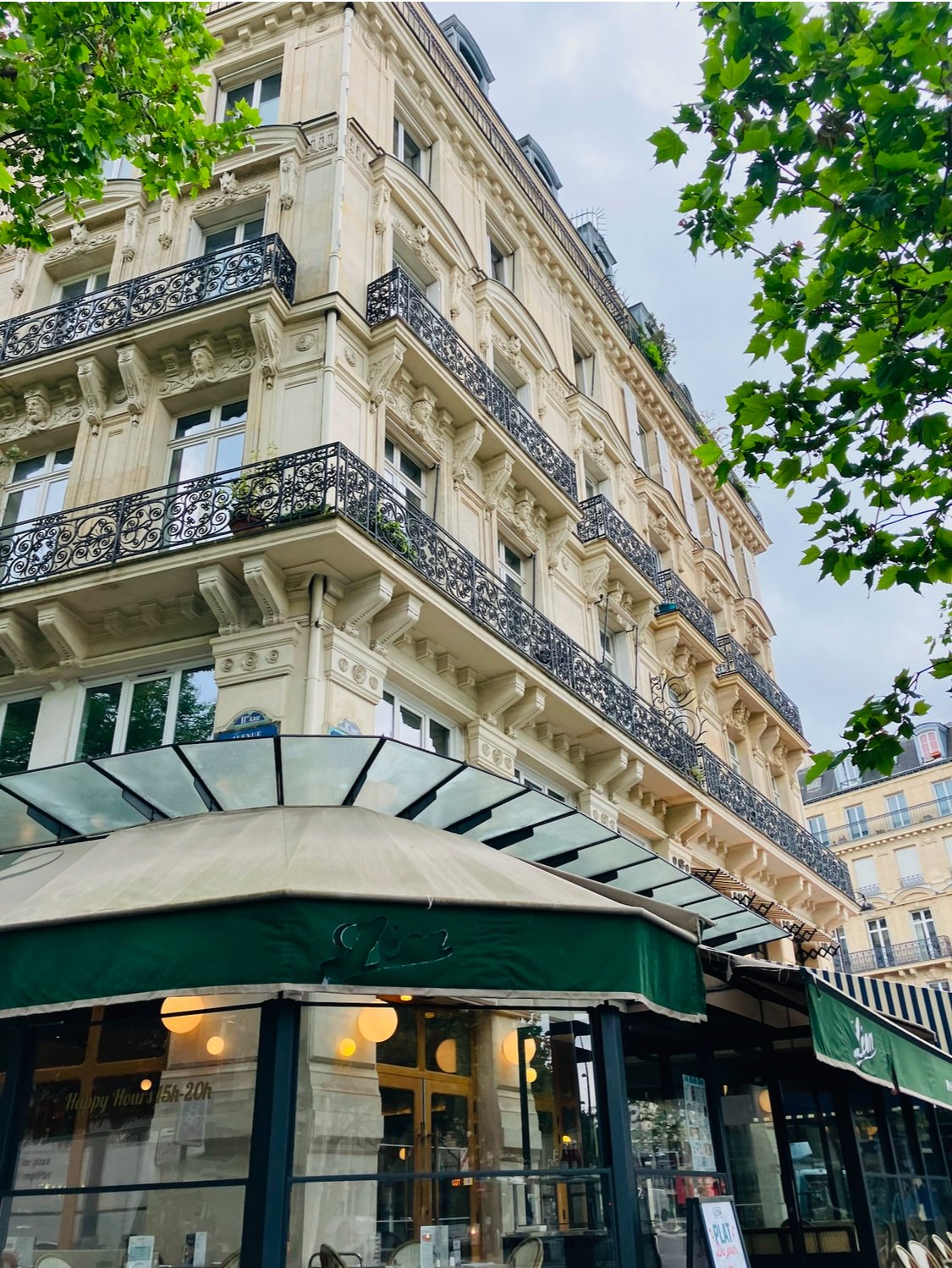 Multi-story Parisian building with ornate balconies, partially obscured by green trees, with a ground-floor café under a green awning.