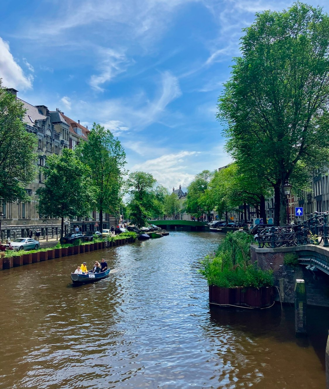 A canal in a European city lined with green trees and classic buildings, with a small boat carrying three people, under a blue sky with a few clouds.
