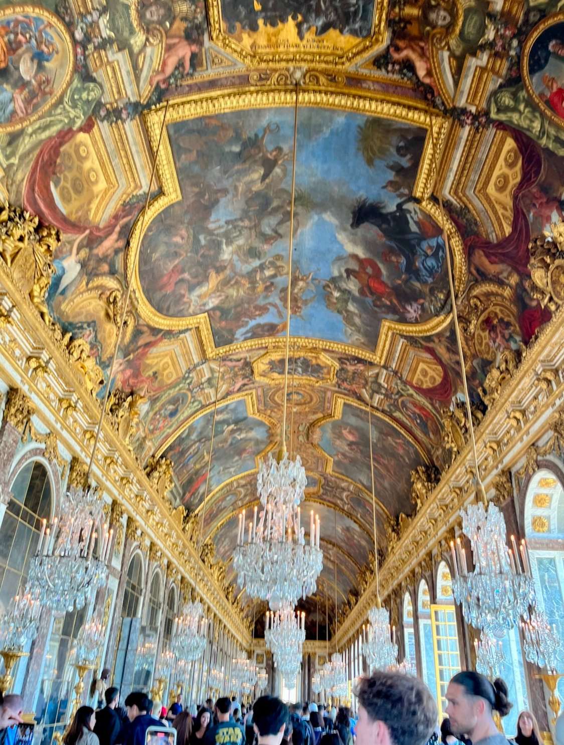 Interior view of the Hall of Mirrors at the Palace of Versailles, featuring a decorated ceiling with paintings, large chandeliers, and many visitors.