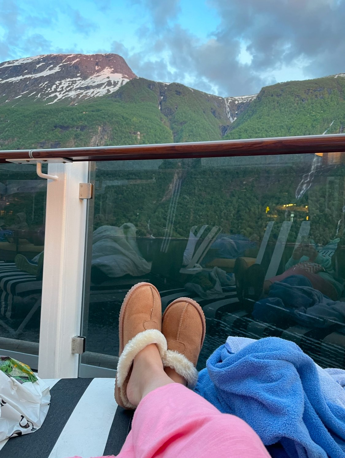 View of a person relaxing on a reclining chair outdoors on a cruise ship, wearing pink pants and tan slippers with a snowy and green mountain in the background.