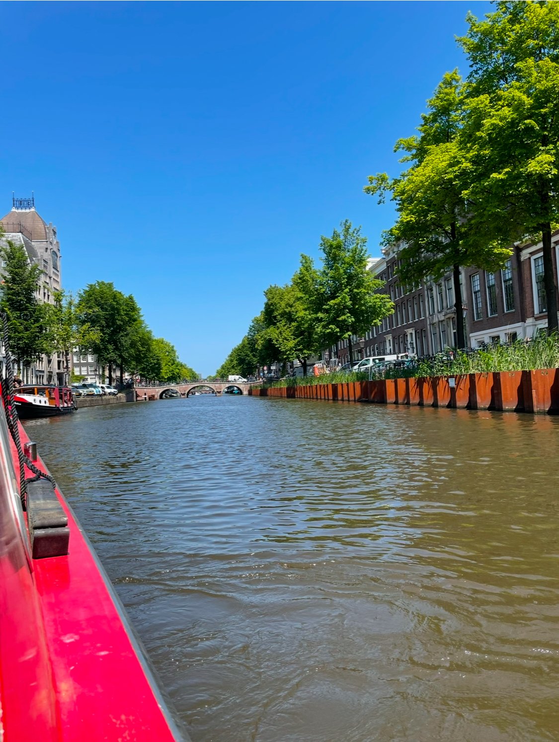 View of a canal with trees on both sides, a red boat in the foreground, and a bridge in the distance under a clear blue sky.