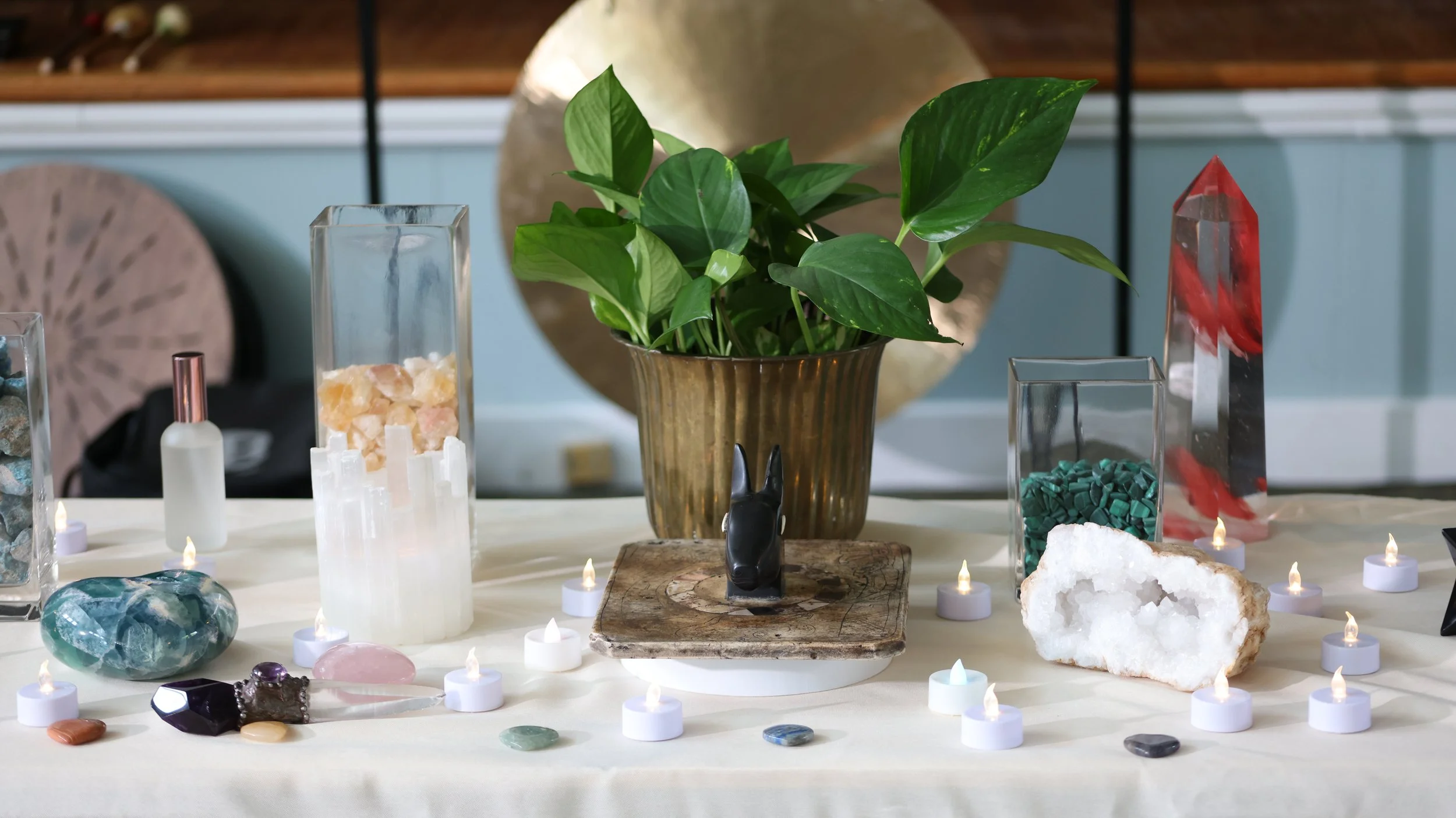 Display of various crystals, gemstones, and decorative objects arranged on a table, including a potted plant, candles, and abstract decorative items.