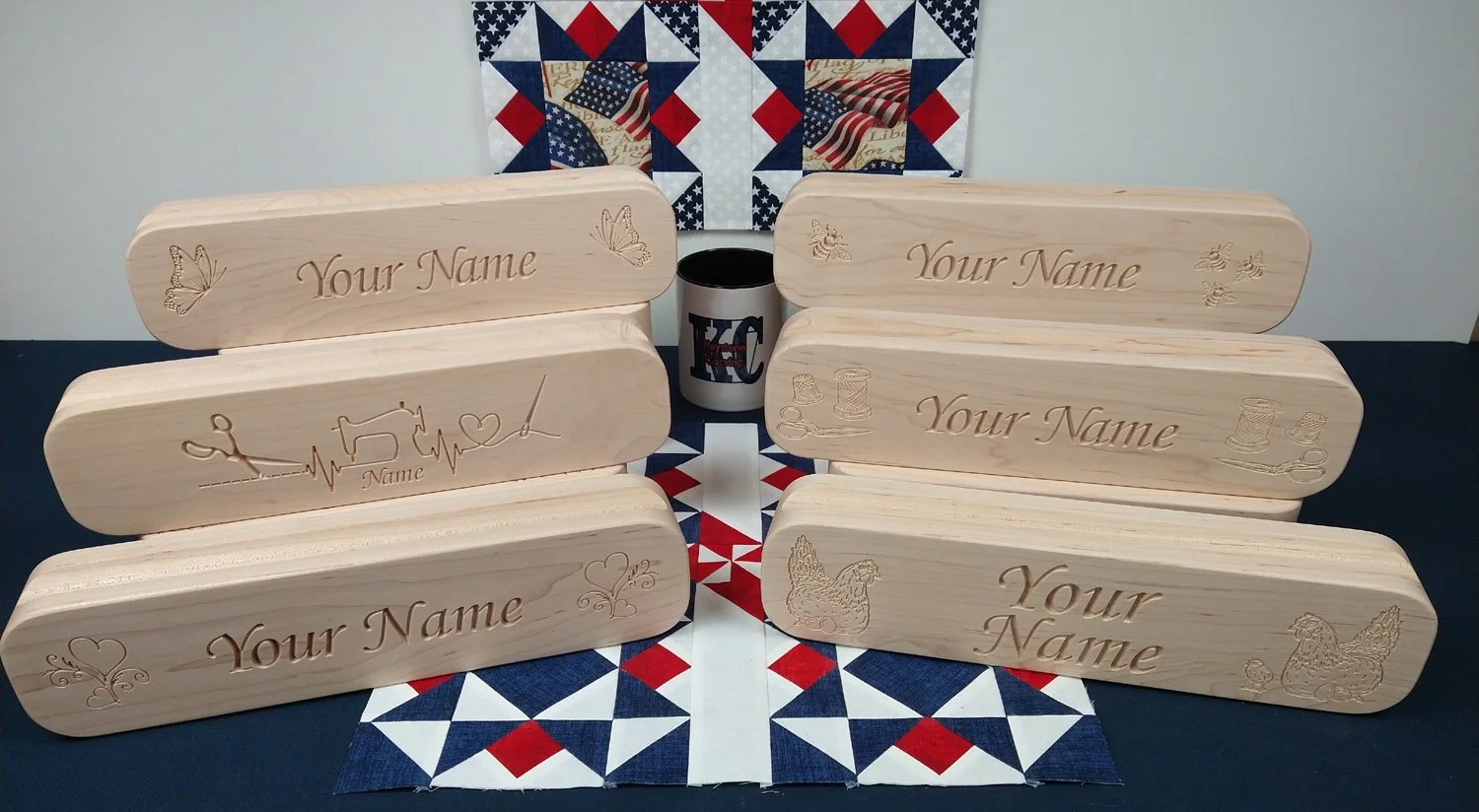Six hard maple quilters clappers, arranged on a table with a patriotic quilt in the background. The clappers feature various personalized wood carvings including butterflies, sewing notions.