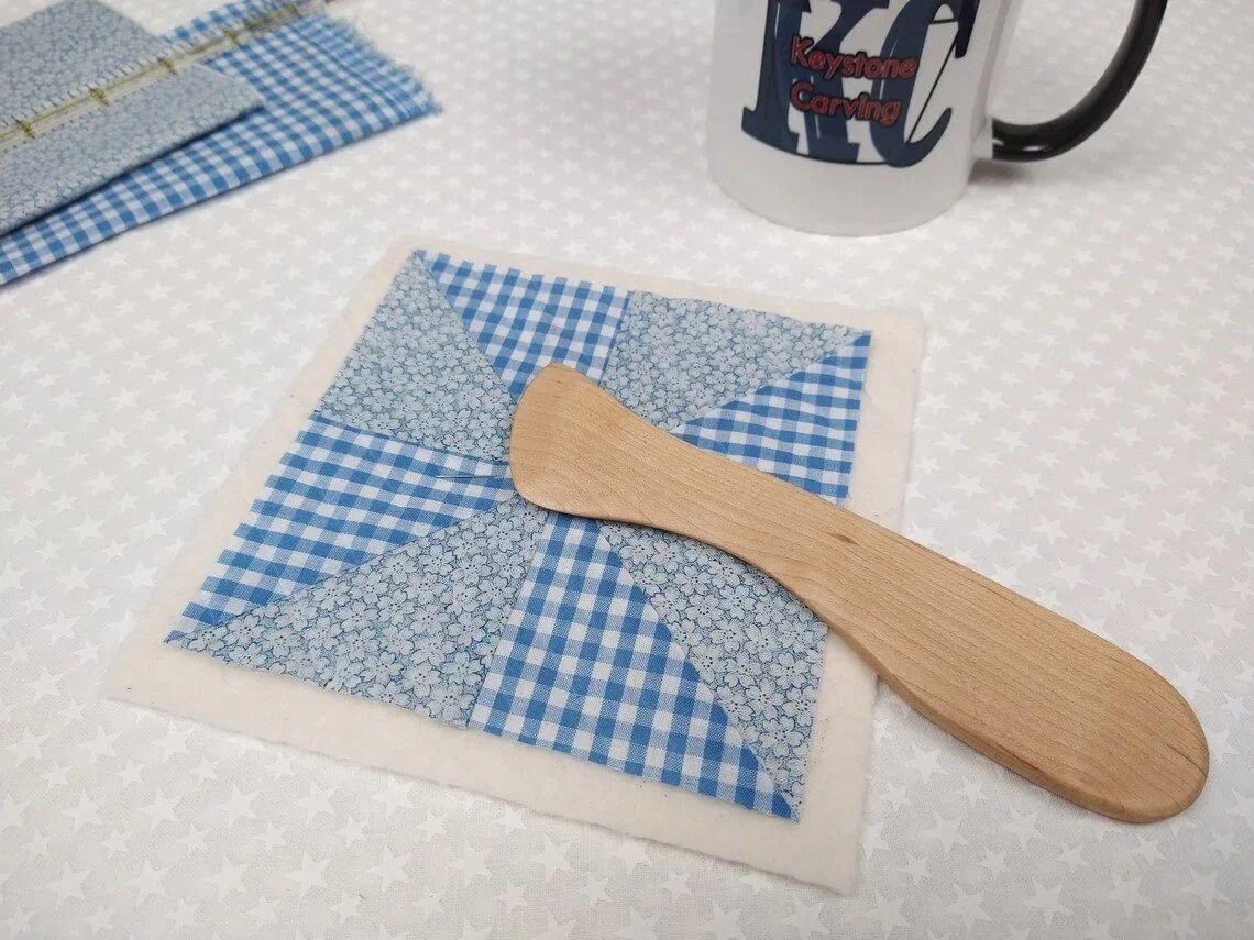Display of a handmade maple Hera marker laying on a blue and white checkered quit block