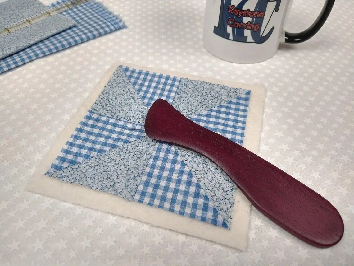 Display of a handmade purple heart Hera marker laying on a blue and white checkered quit block
