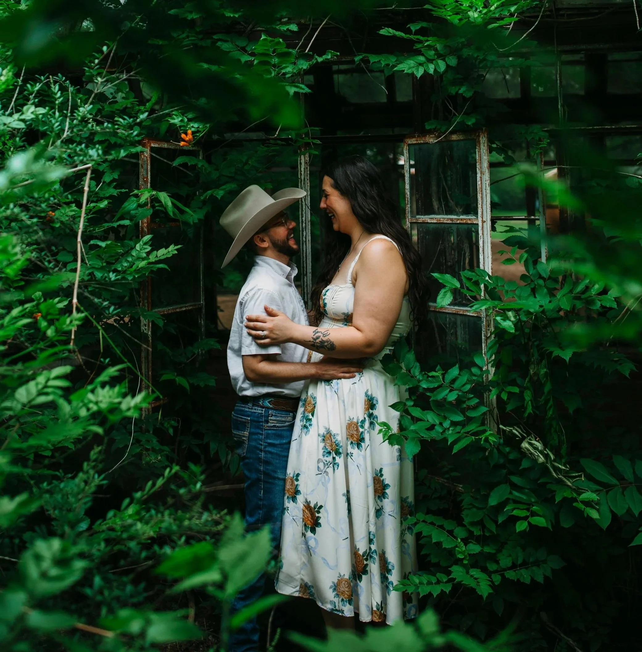 Miriam and her husband stand close together in a lush, green outdoor setting surrounded by dense foliage. They are smiling and looking at each other, with the woman in a white floral dress and the man in a light-colored shirt and a hat.