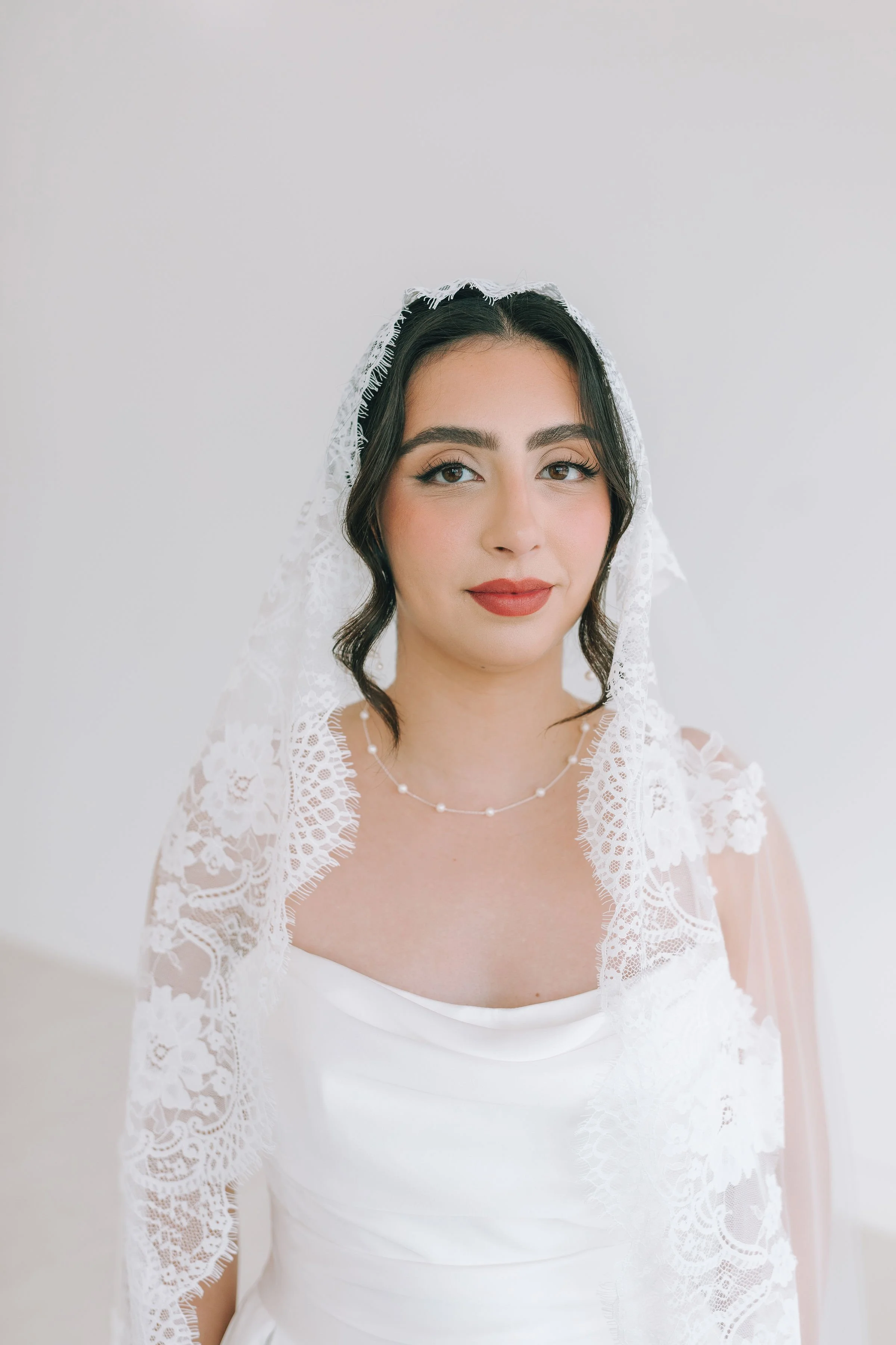 A woman in a white wedding dress with lace details and a lace veil looks slightly to her right, standing against a plain, light-colored background.