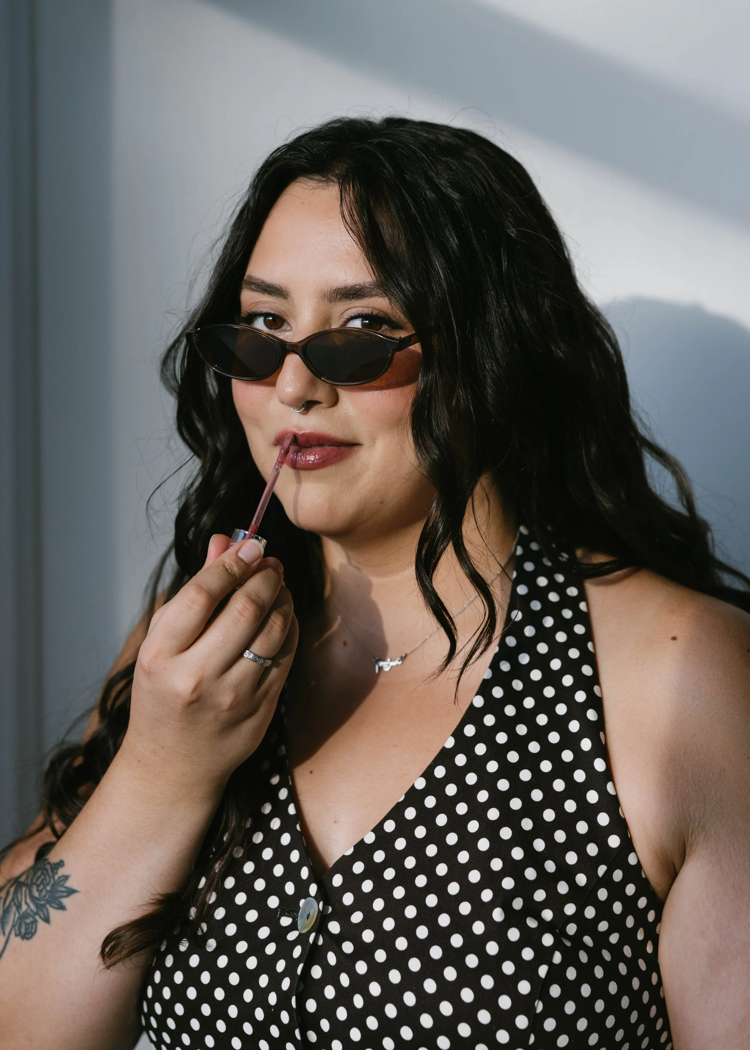 Miriam, a woman and the owner of MBM Co, with dark hair wearing black sunglasses, applying lip gloss with a polka dot shirt.